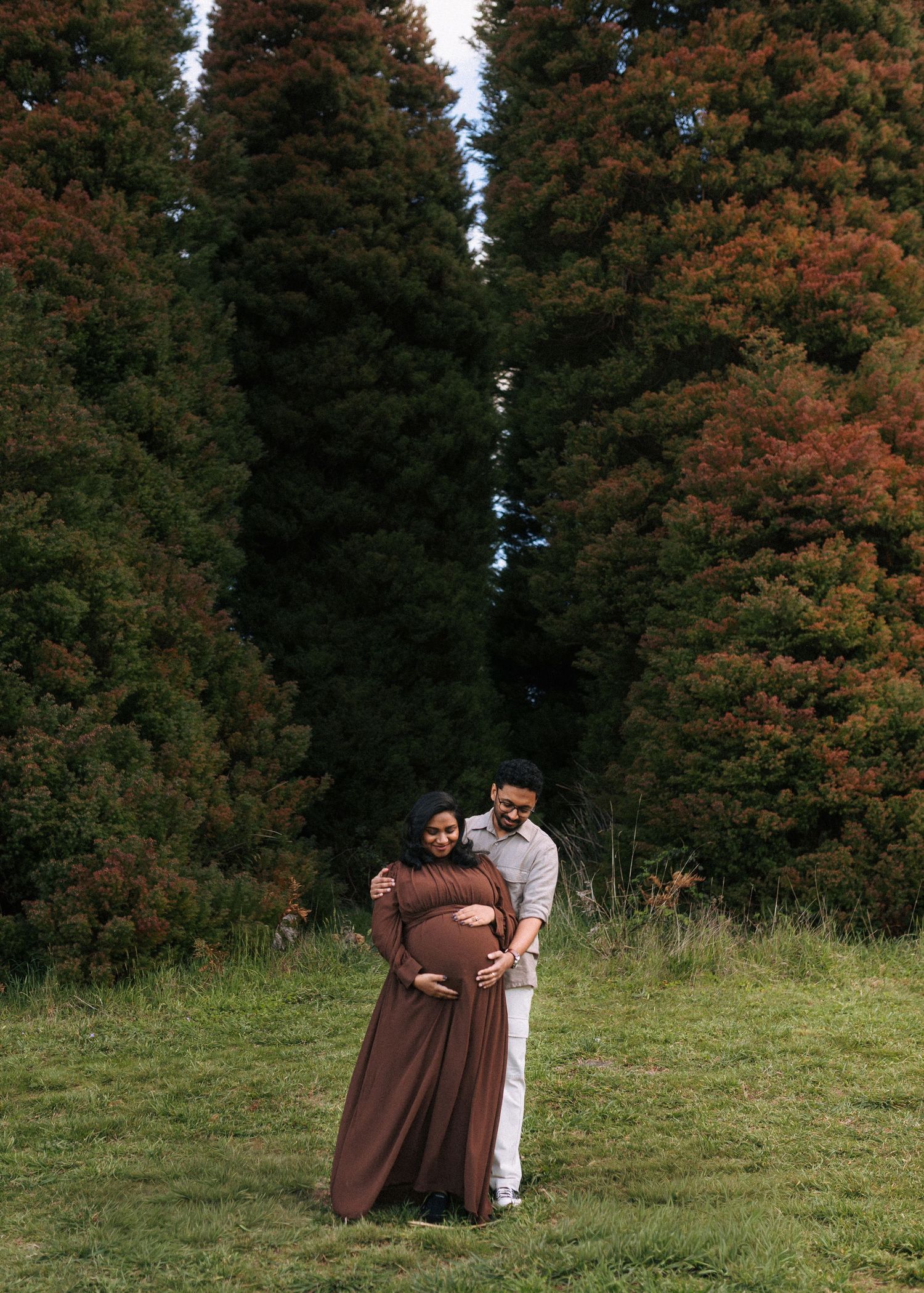 Expecting couple embracing in a grassy field during a maternity photoshoot, mother-to-be in a brown dress, with tall evergreen trees behind.