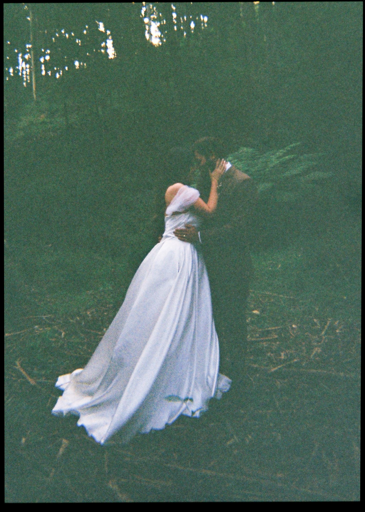Bride in flowing white off-shoulder gown standing gracefully among ferns in a lush dark forest setting.