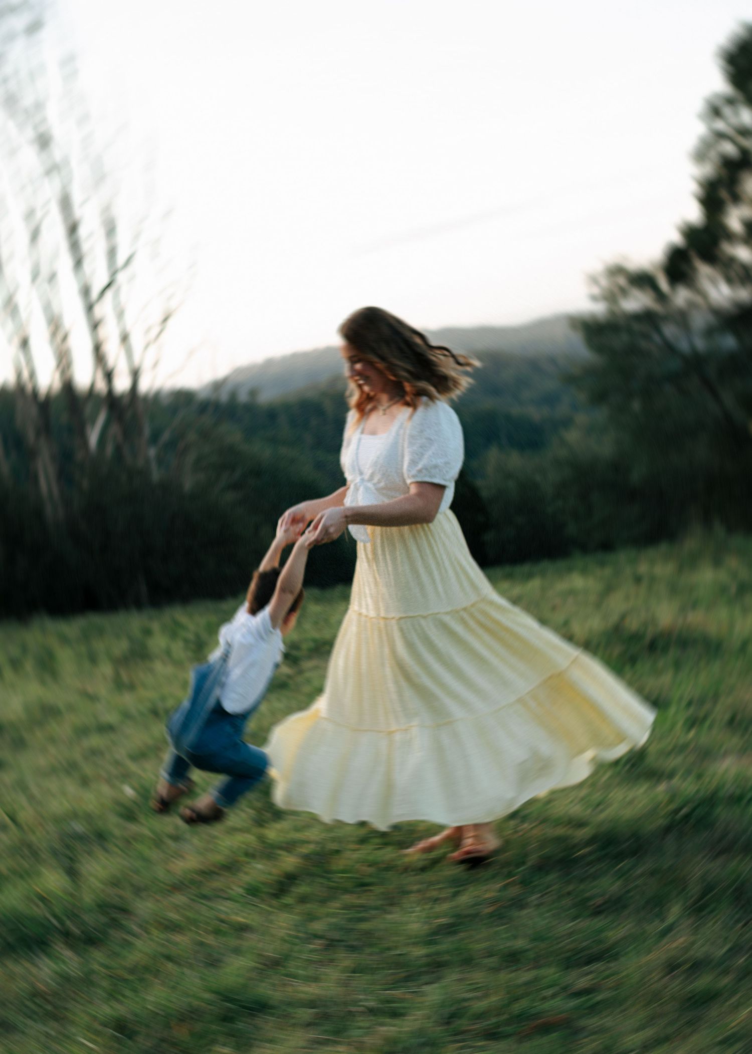 A mother in a yellow skirt twirls her laughing toddler son in a sunny outdoor meadow.