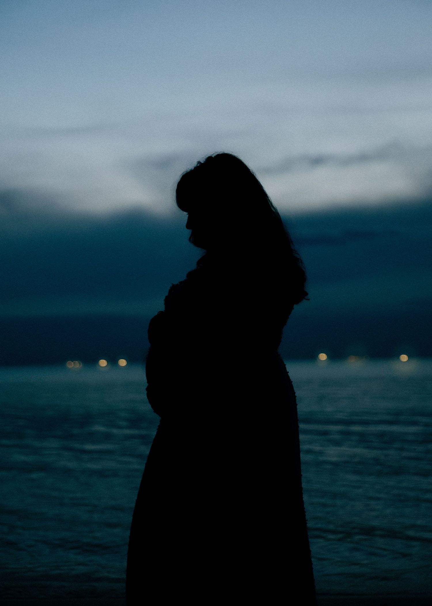 Silhouette of a pregnant woman holding her baby bump by the ocean at dusk.