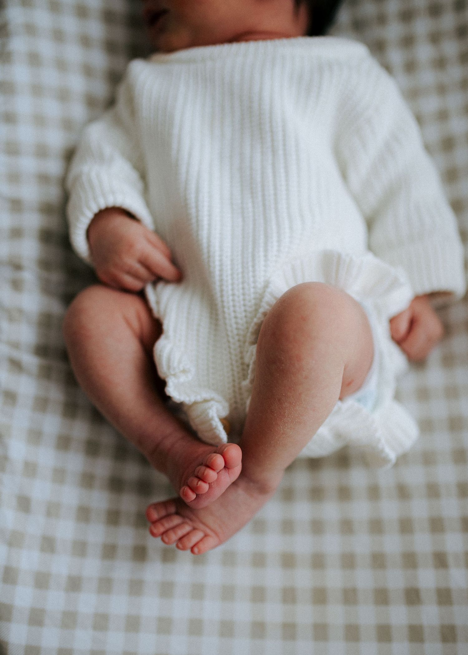 Detail shot of newborn baby legs and feet in white knit romper with ruffle trim on gingham bedding.