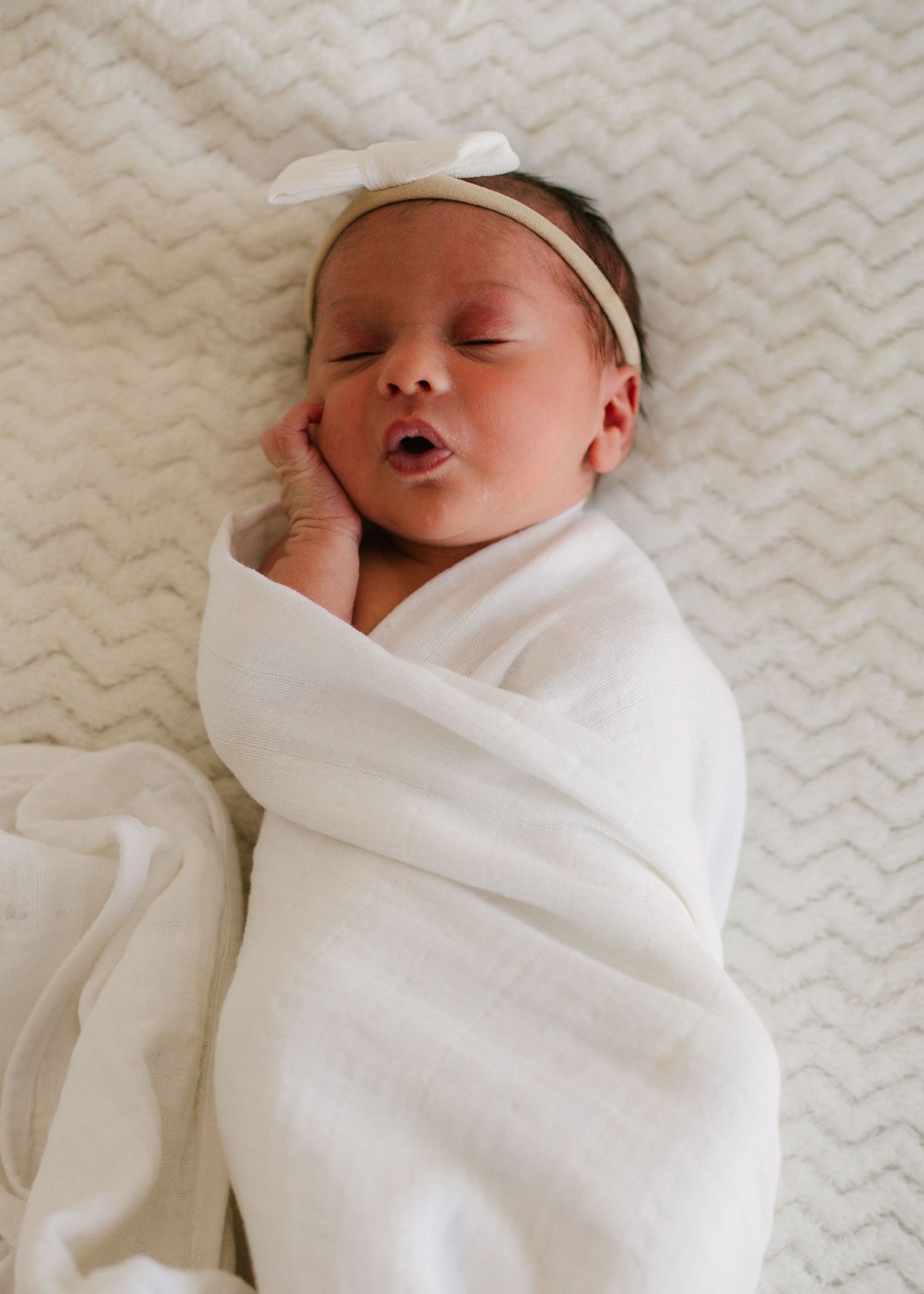 Peaceful newborn baby swaddled in white blanket with bow headband sleeping with mouth open.