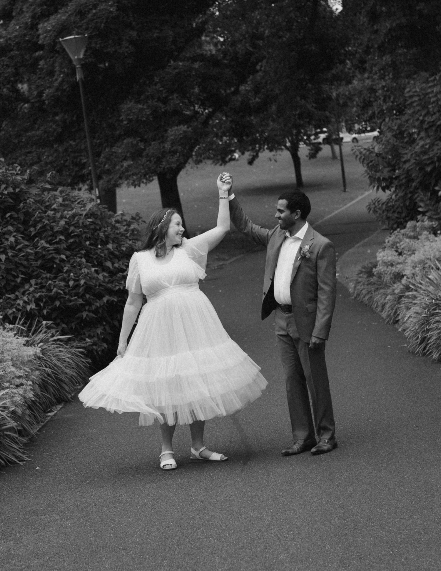 Black and white photo of bride and groom dancing on a park path, groom spinning bride in white tulle dress.