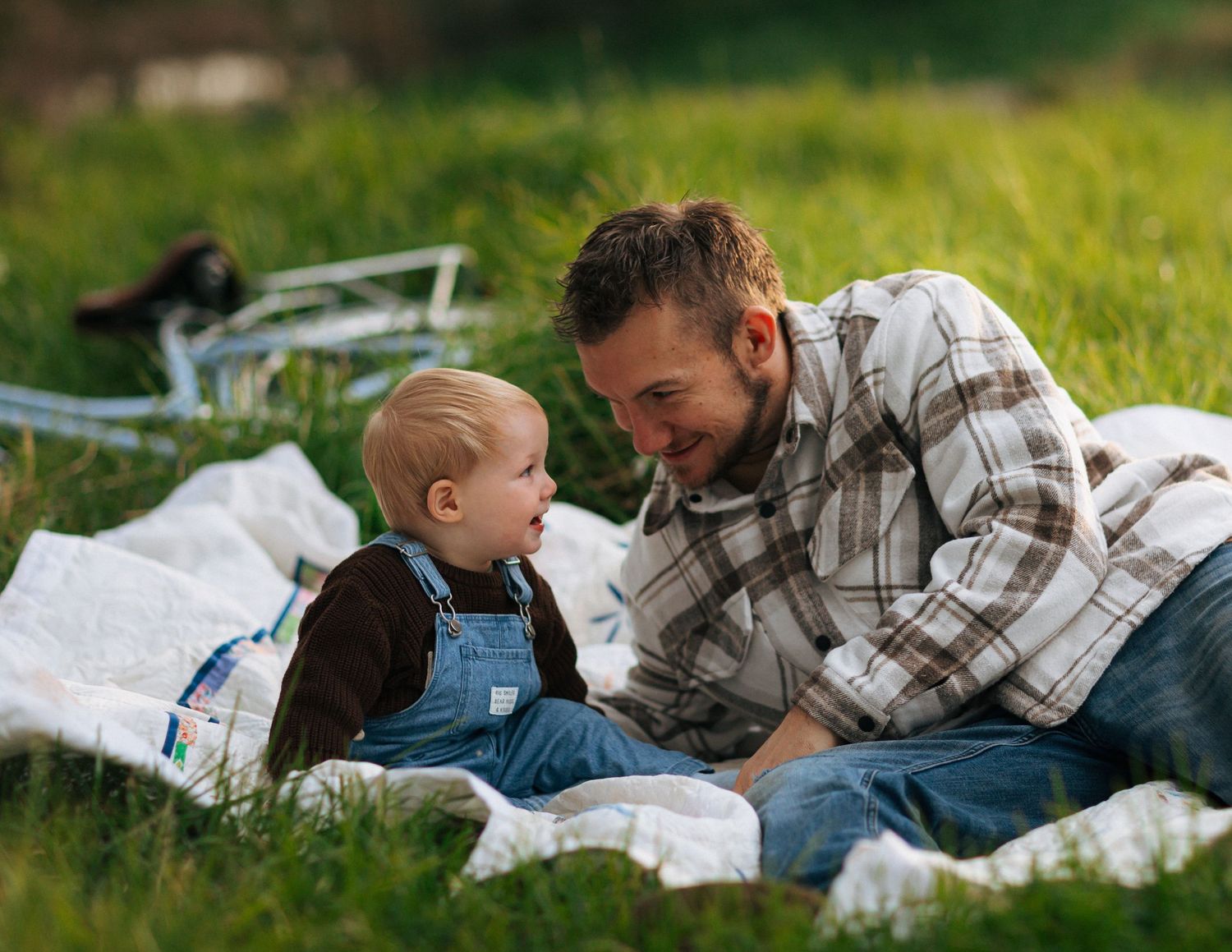 Father and toddler sharing a sweet moment together on a blanket in sunny green grass outdoors.
