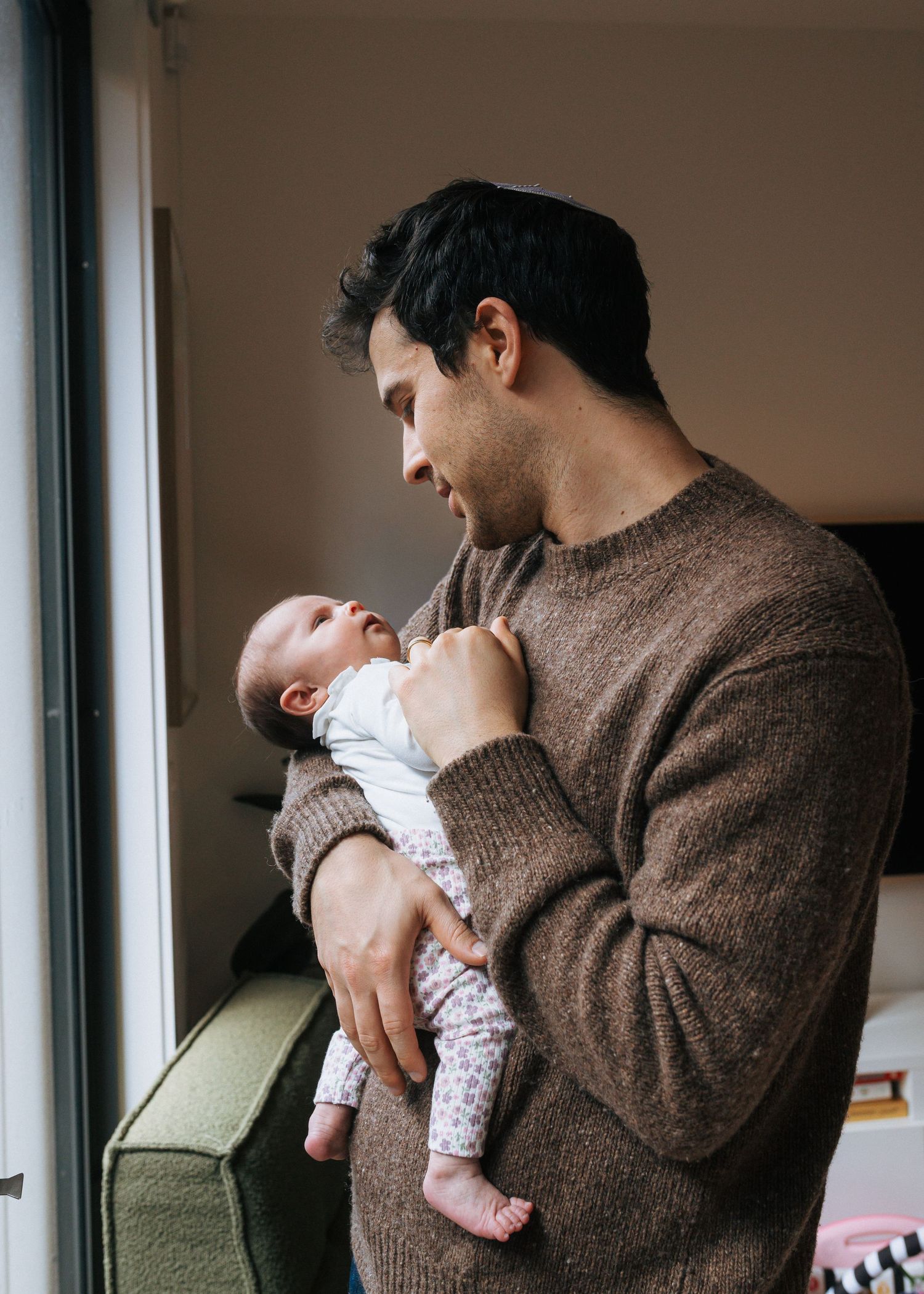 New father in brown sweater tenderly holding and looking at his newborn baby by window.