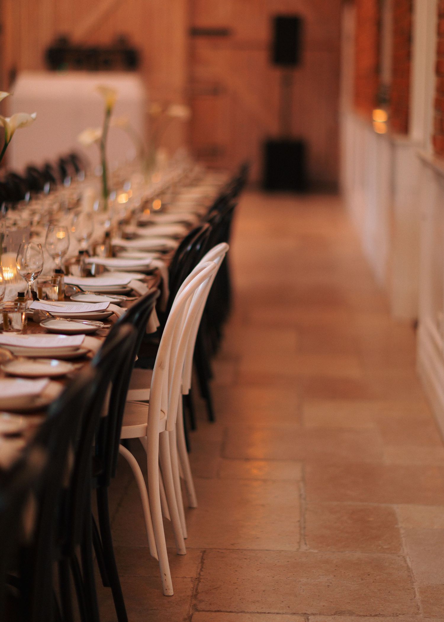 Elegant long reception table with white chairs, candles, and place settings in a warmly lit venue.