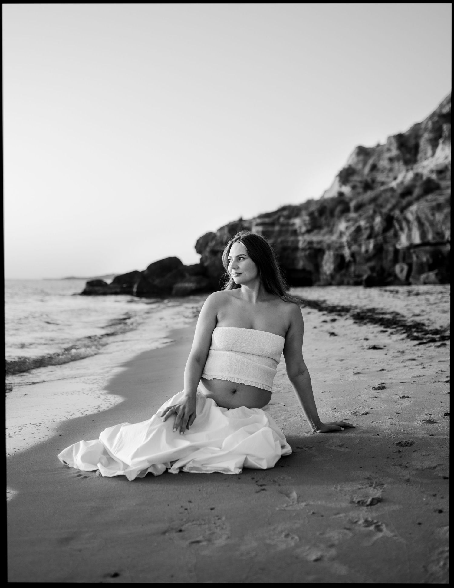Black and white photo of a pregnant woman sitting gracefully on a sandy beach near rocky cliffs.