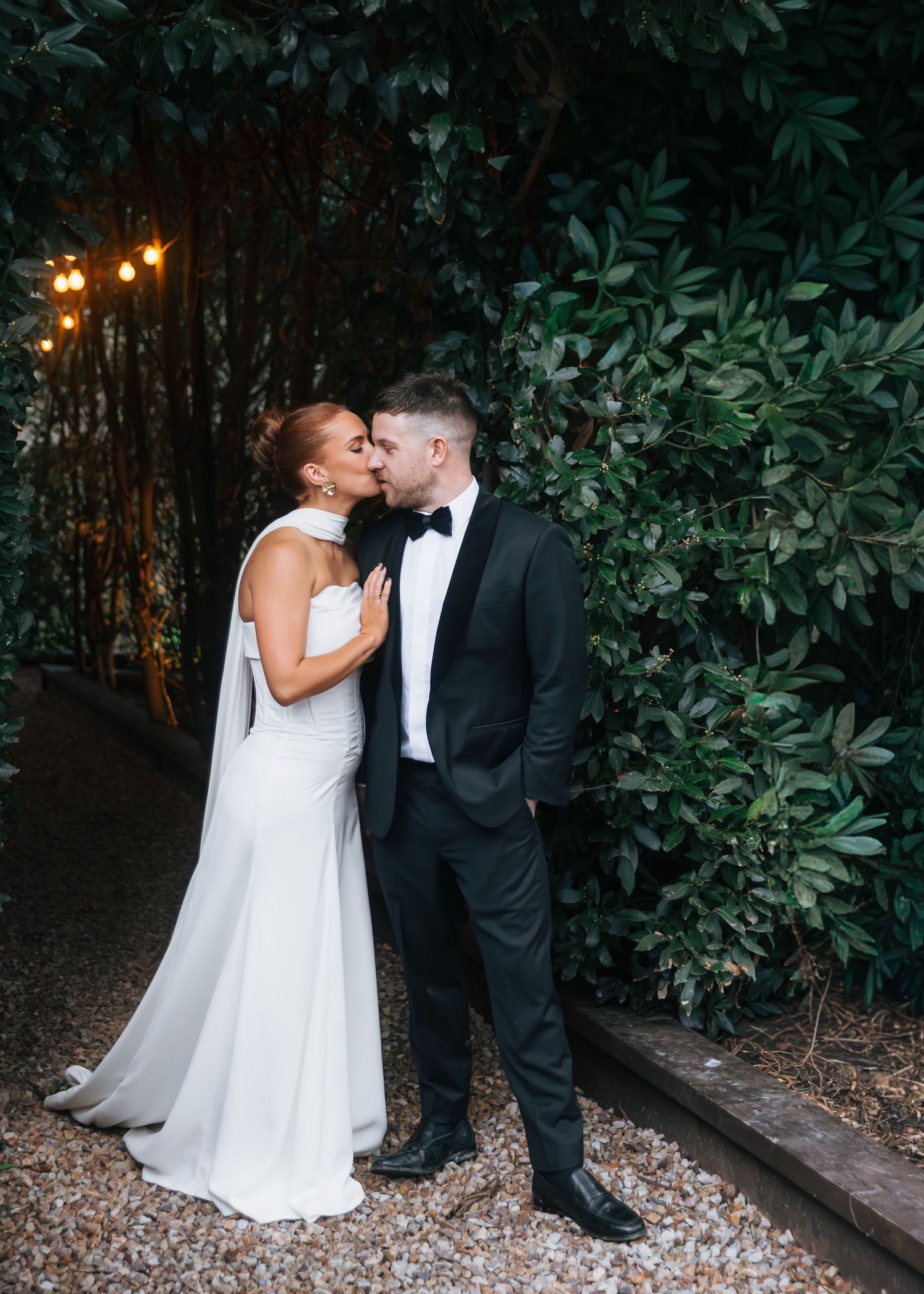 Bride in white gown kisses groom in black tuxedo amid lush green foliage with warm string lights.