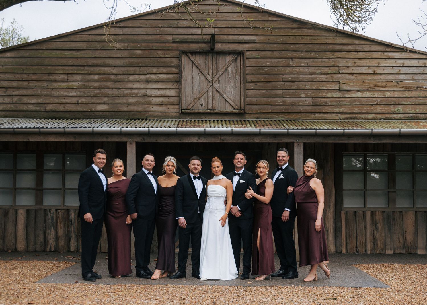 Wedding party poses in burgundy dresses and black tuxedos before a rustic barn venue.