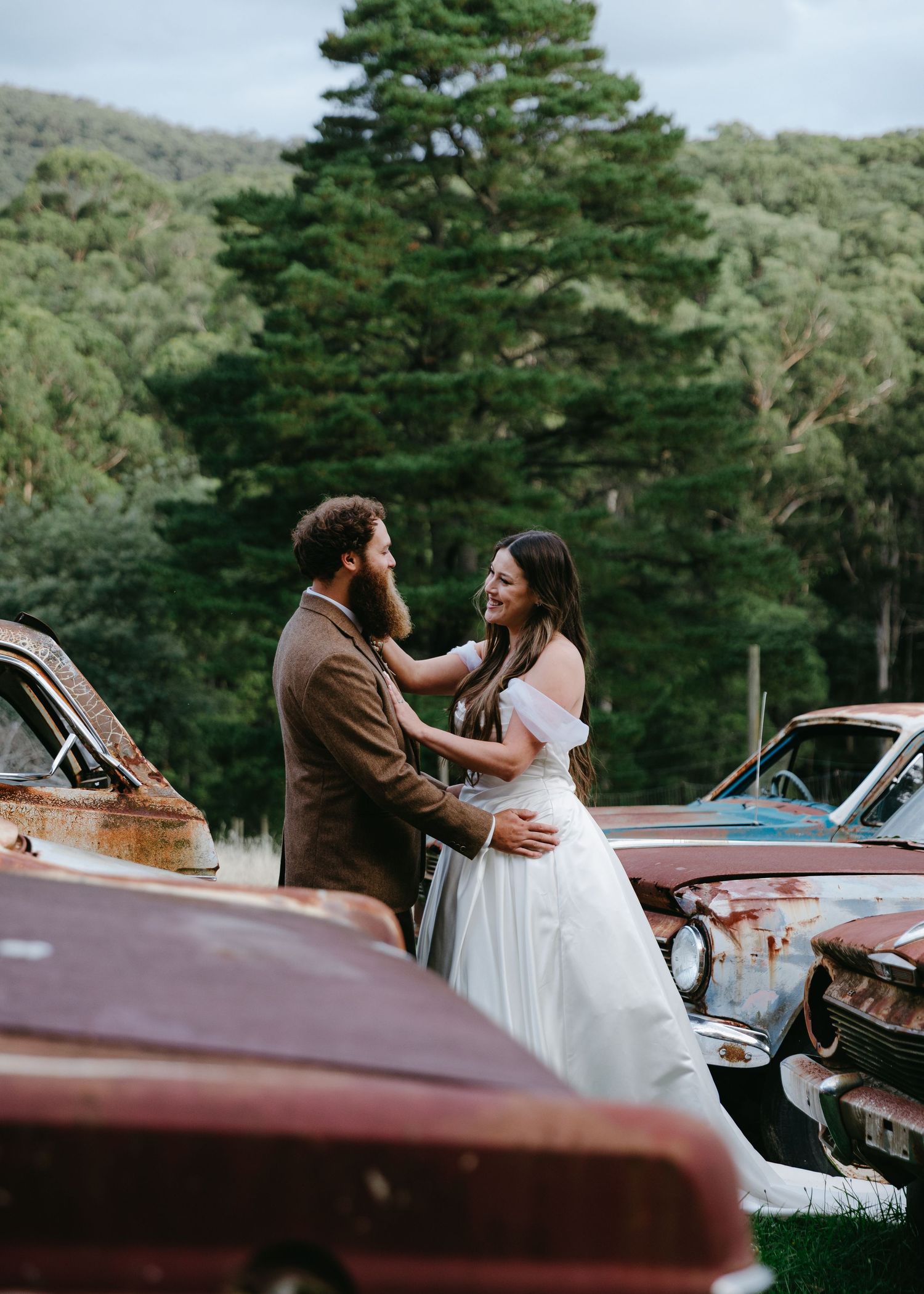 Bride and groom sharing a joyful moment surrounded by vintage rusty cars in a scenic outdoor setting.