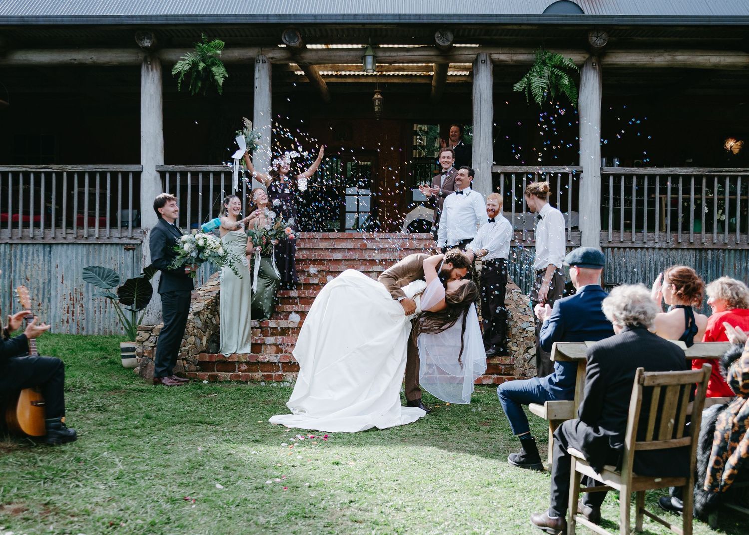 Groom dipping bride for a kiss as guests celebrate with confetti on rustic venue steps after ceremony.