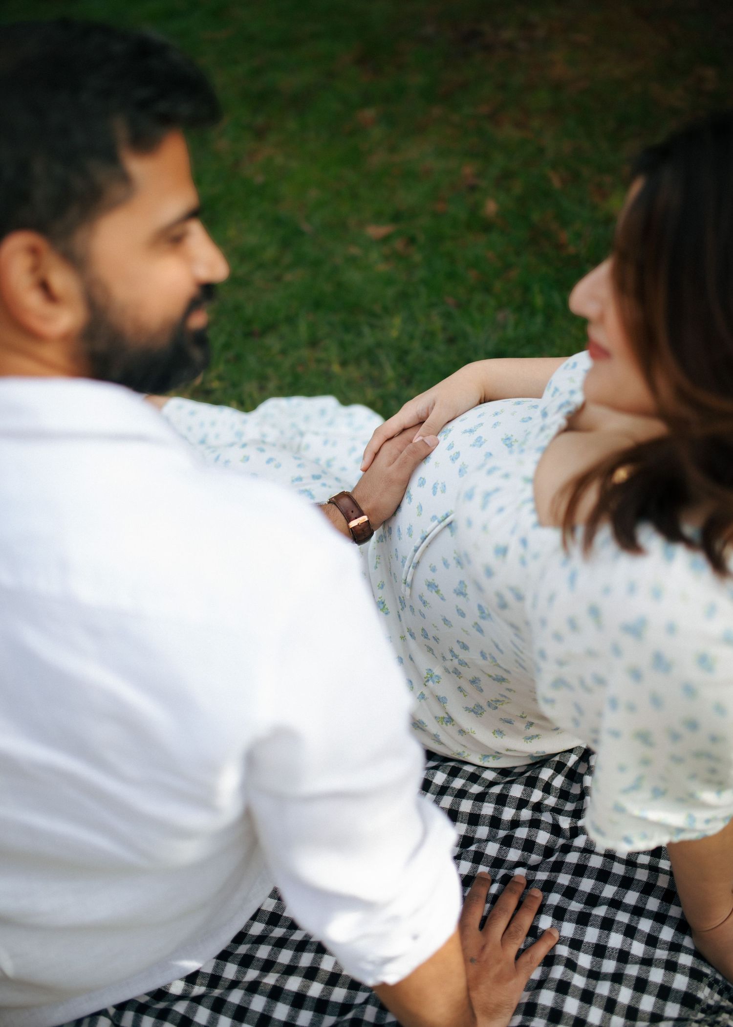 Couple sitting on a gingham picnic blanket outdoors, man gently touching pregnant woman's baby bump.