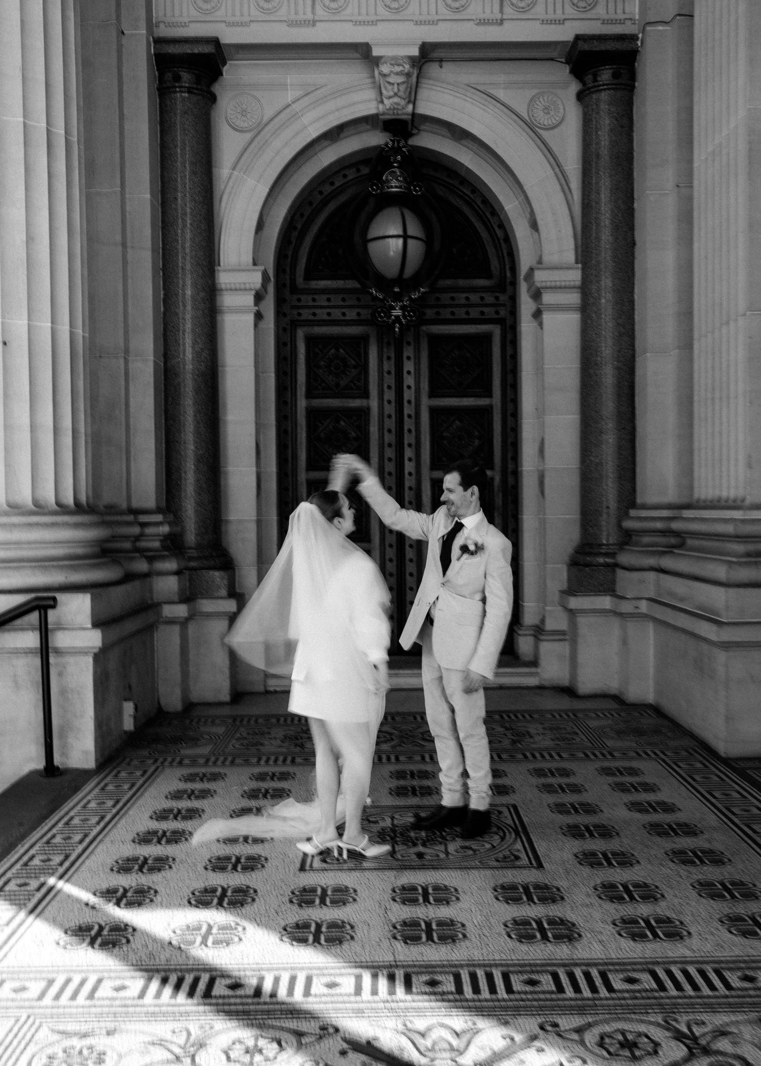 Black and white photo of groom spinning bride in short dress and veil at grand building entrance.