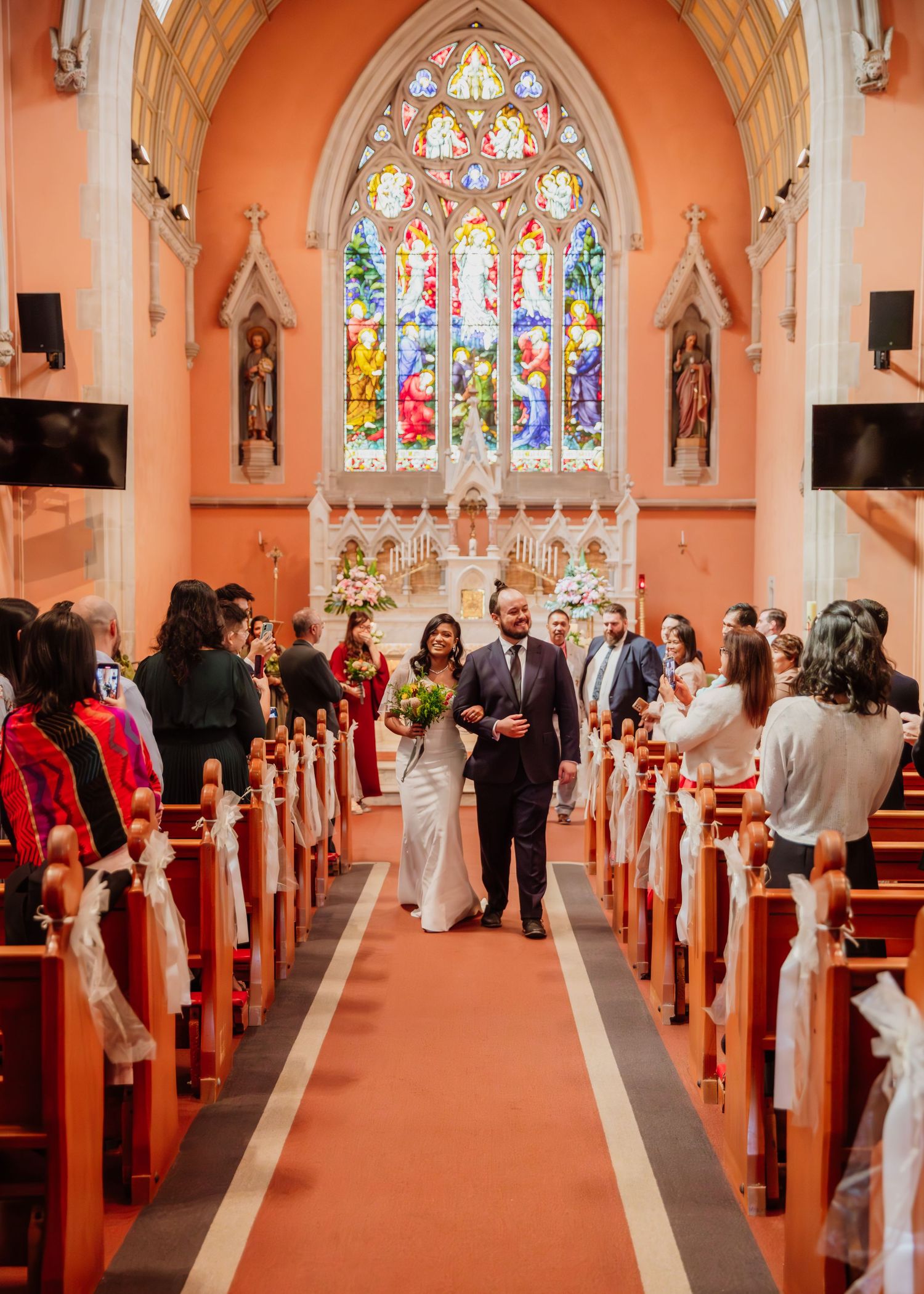 Newlyweds walk down the aisle of a beautiful church with stained glass windows after their wedding ceremony.