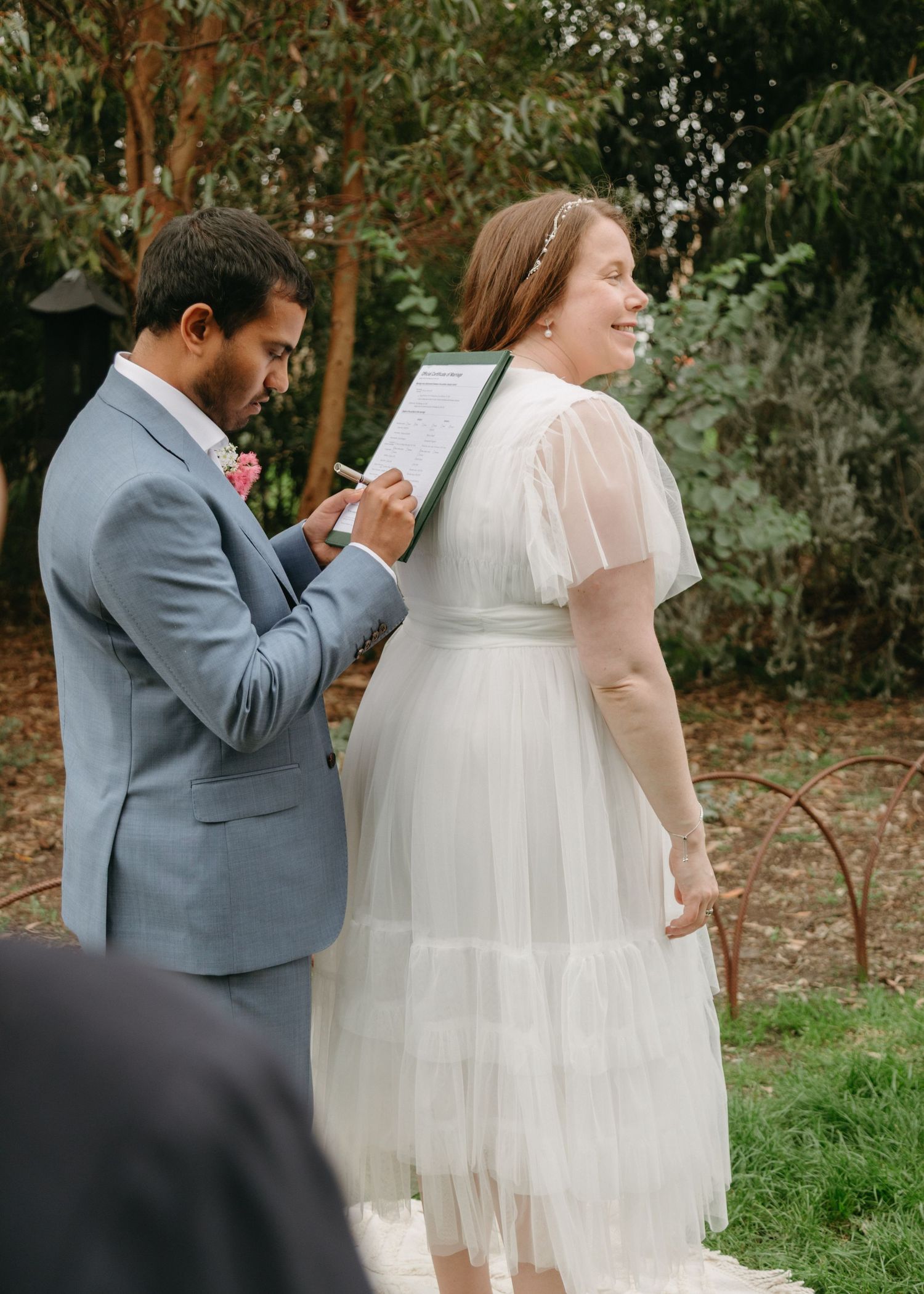 Groom in grey suit signing marriage certificate while smiling bride stands beside him outdoors.