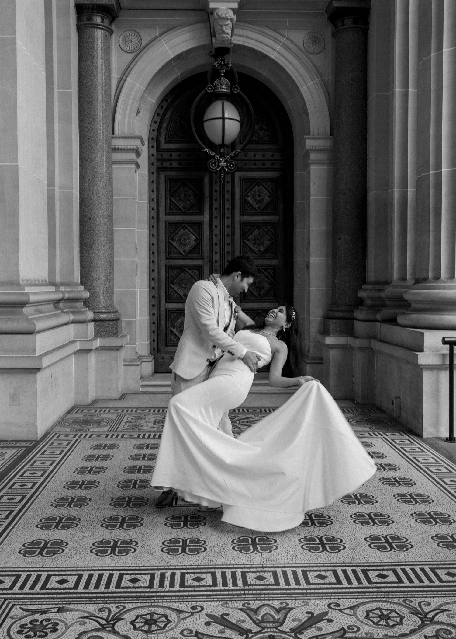 Black and white photo of groom dipping bride in romantic pose before ornate historic building.