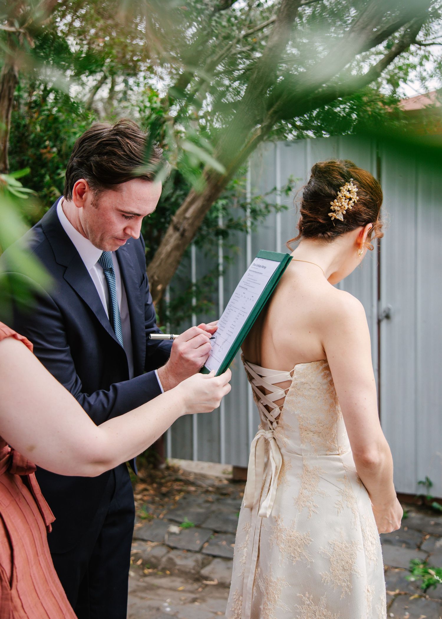 Groom signing wedding documents on clipboard while bride stands nearby in garden.