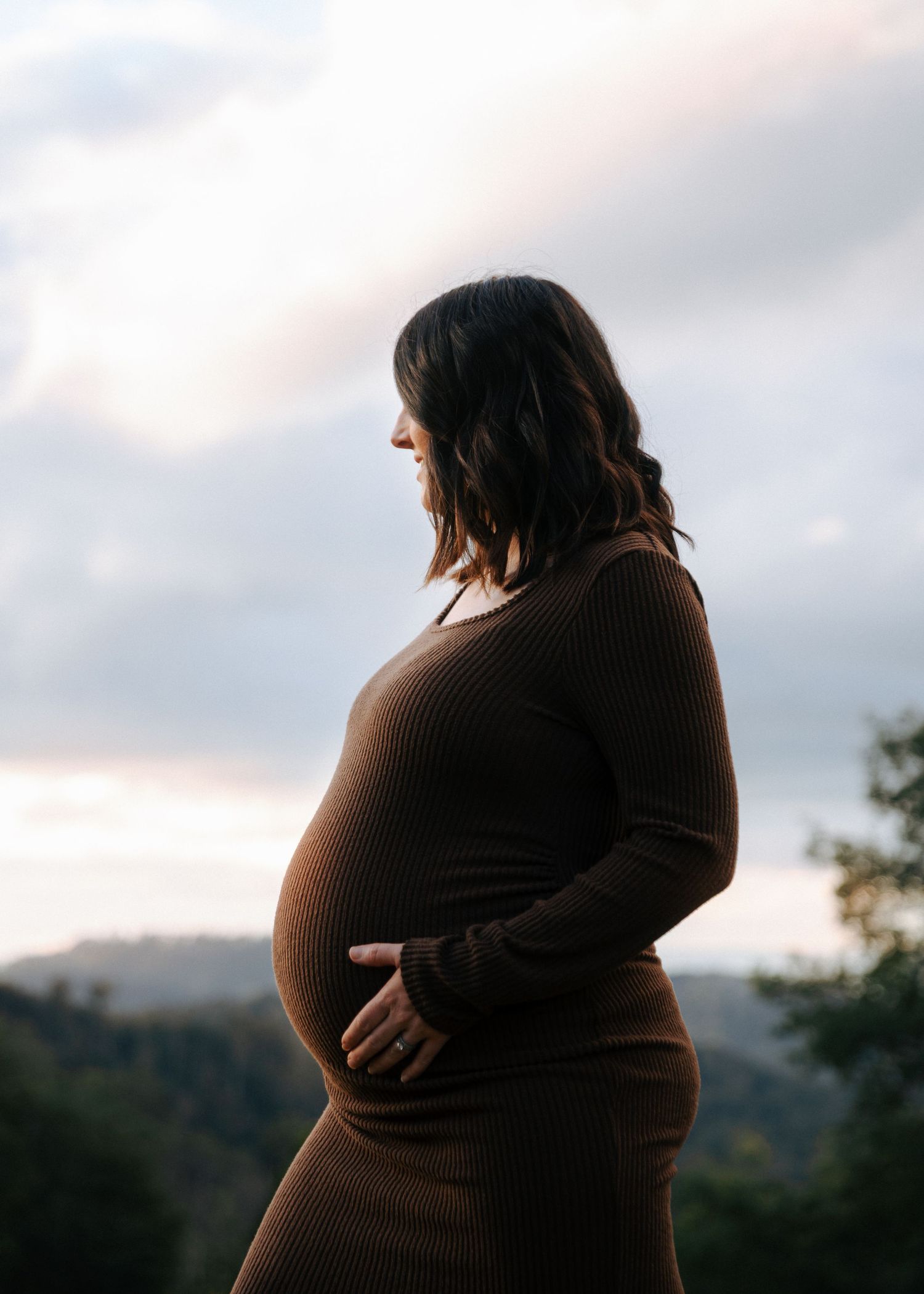 Side profile of pregnant woman in brown ribbed dress holding bump at dusk.