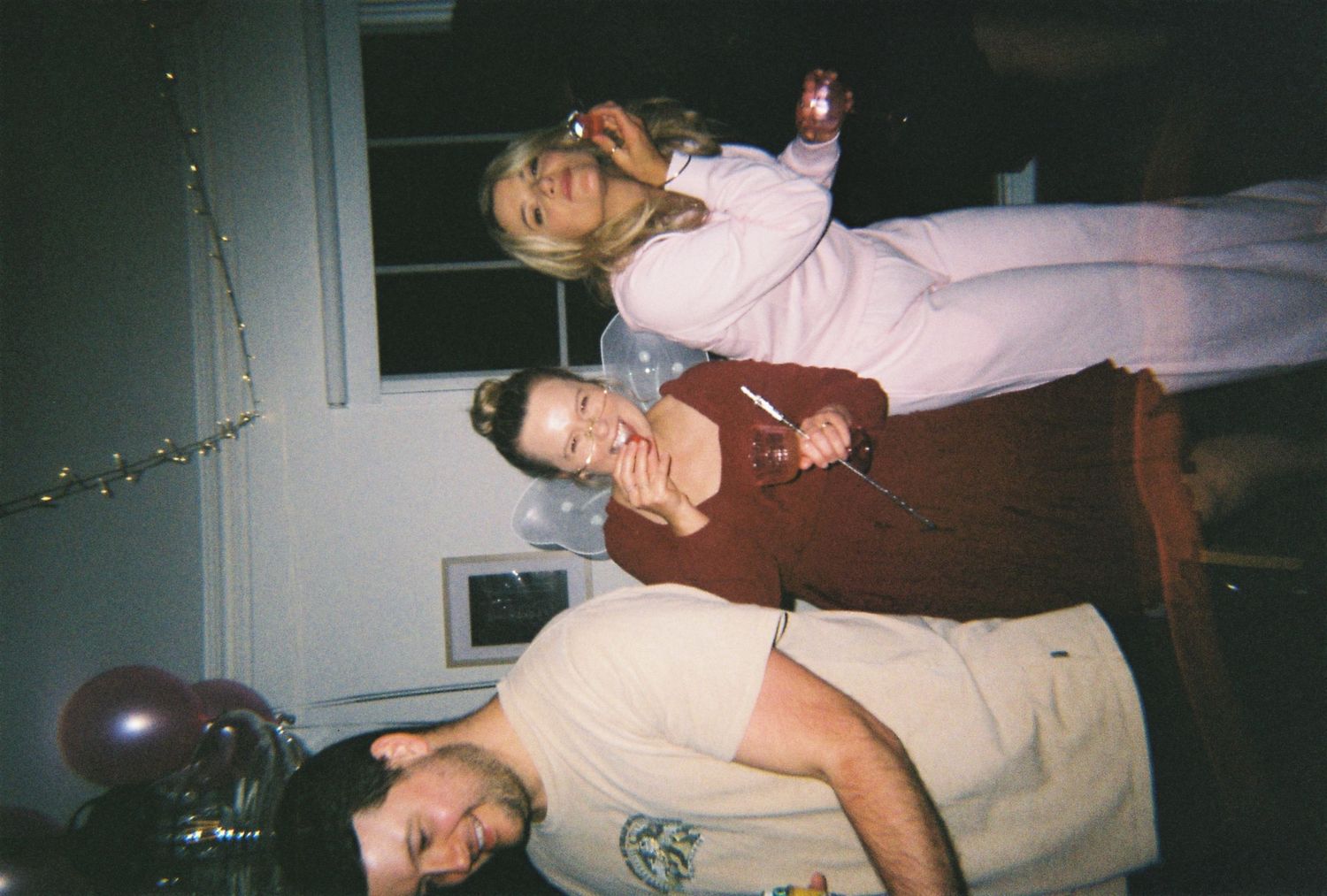 Three people at a party, with a woman in pink standing behind two men, balloons visible in background.