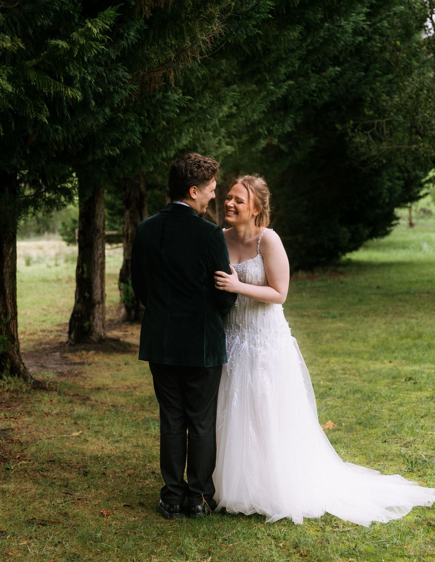 Smiling bride in a white lace wedding gown embraces a groom in a dark velvet jacket on a grassy lawn beneath tall evergreen trees.