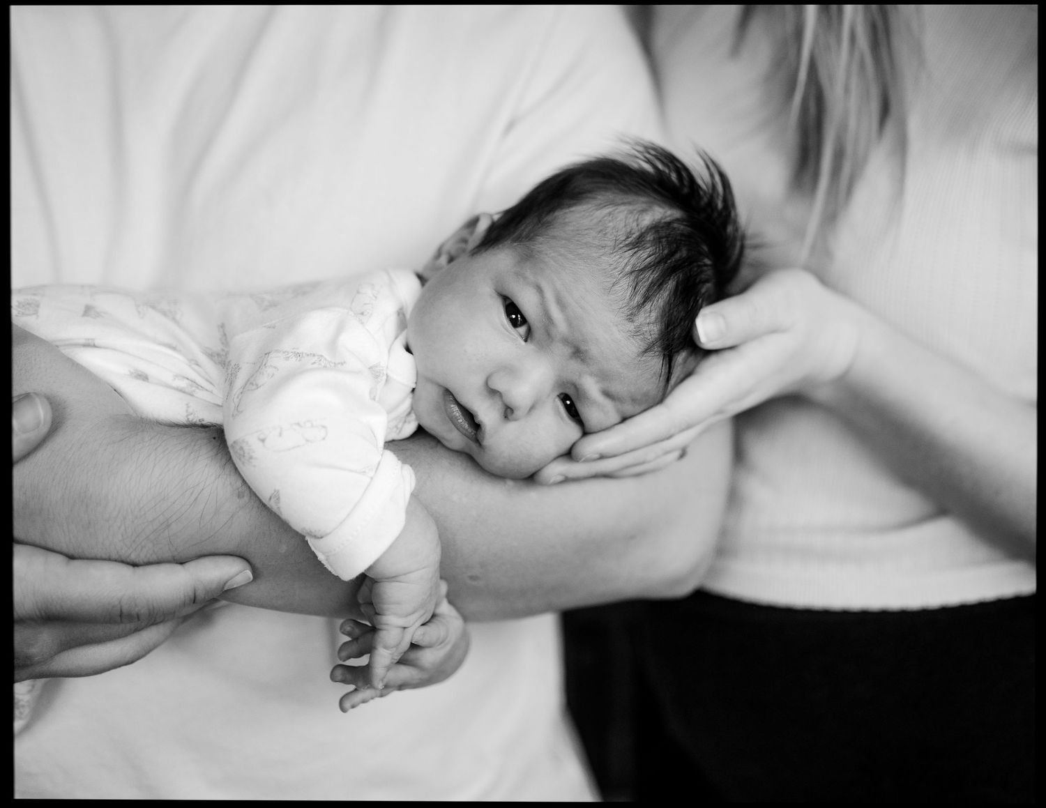 Black and white photo of a newborn baby with dark hair resting in a parent's arms while another hand gently cradles the baby's head.