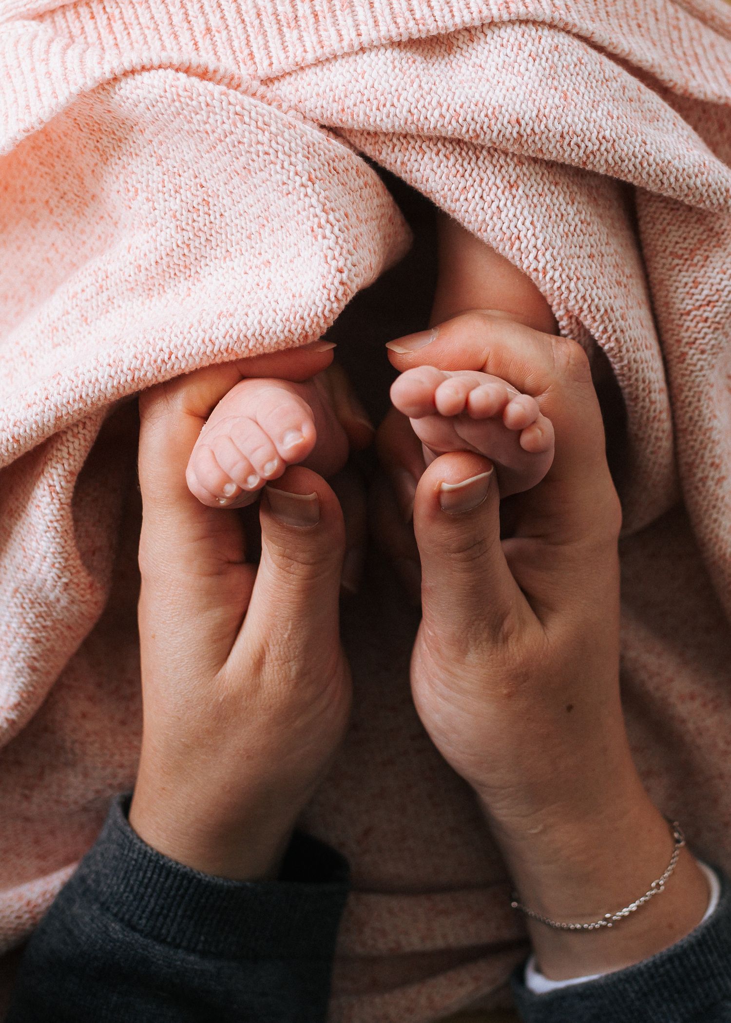 Two adult hands gently cradle a newborn baby's tiny feet wrapped in a soft pink knit blanket.