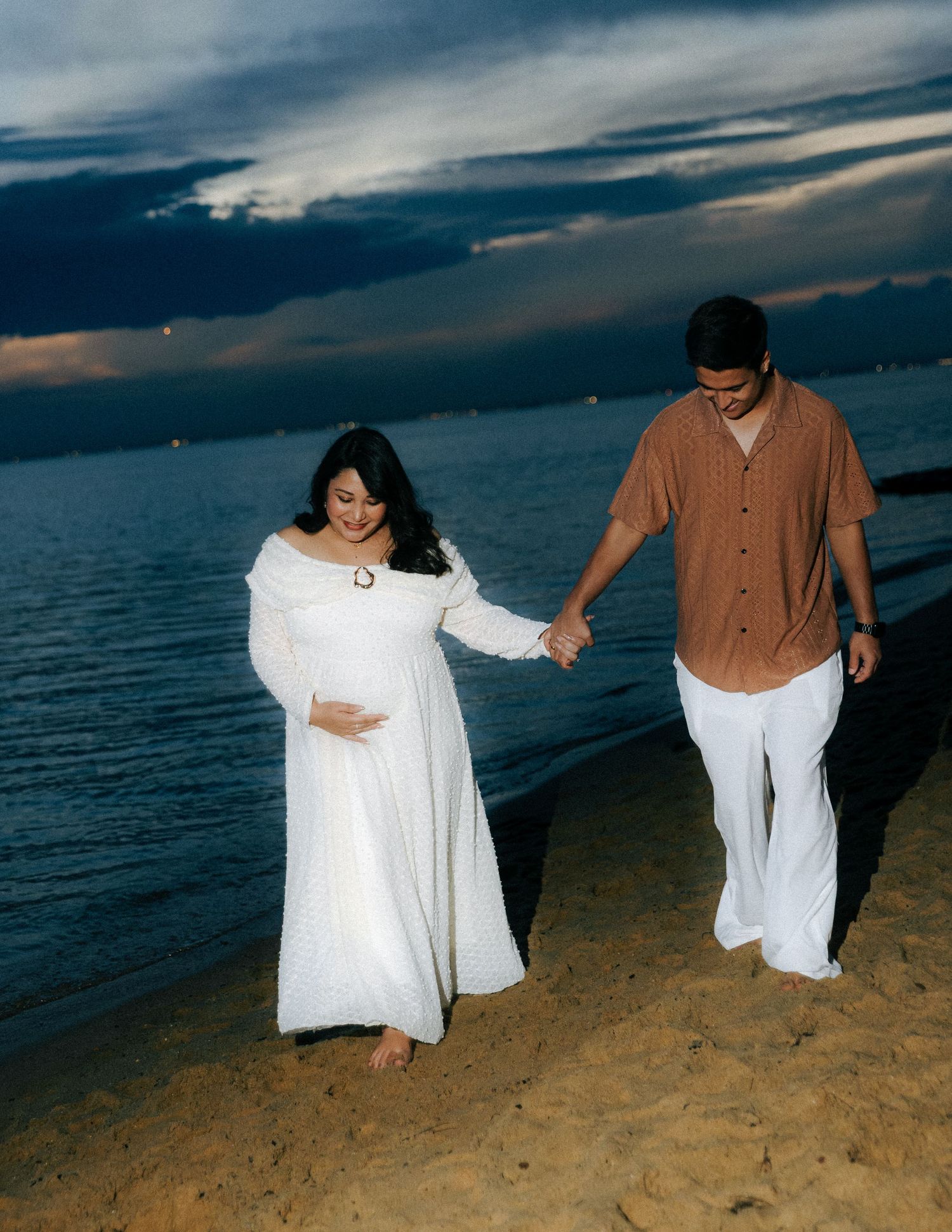 Pregnant couple holding hands walking on sandy beach at sunset near the ocean.