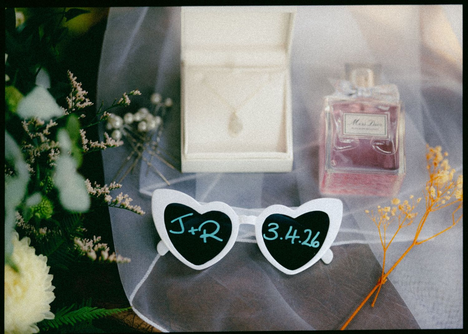 Wedding flat lay featuring heart-shaped sunglasses with J+R 3.4.26, Dior perfume, and floral veil.