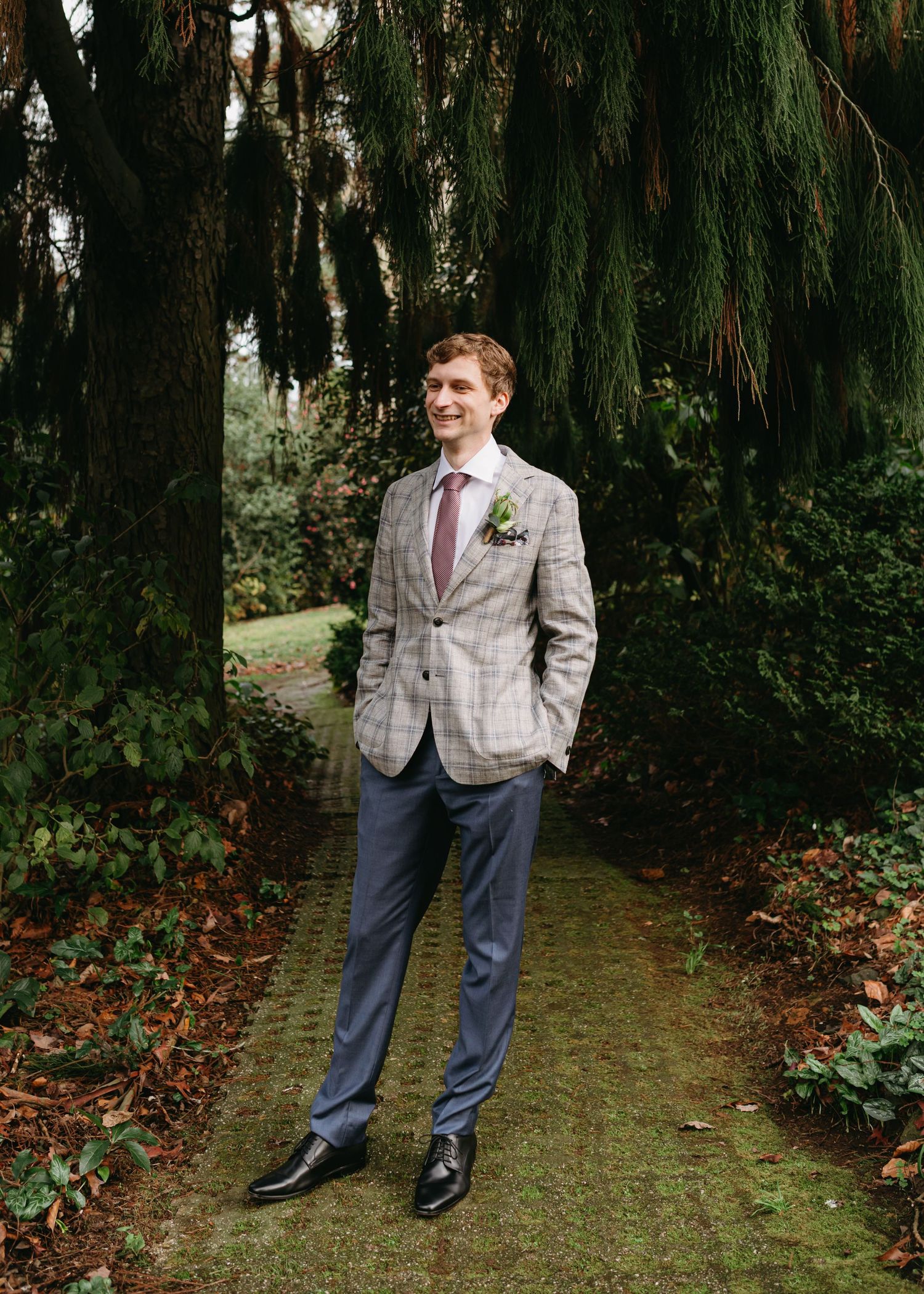Groom in a grey plaid blazer and blue trousers standing on a garden path.