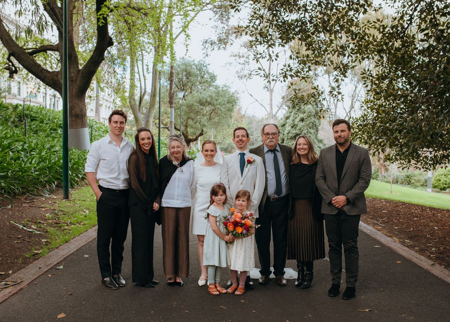 Family wedding portrait on scenic park pathway with bride, groom, and loved ones smiling together.