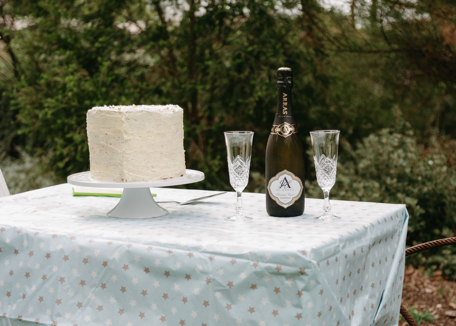 White wedding cake displayed with champagne bottle and crystal flutes on outdoor table.