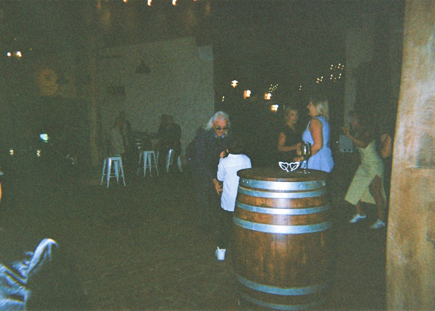 Wedding reception guests socializing around a wooden barrel in a dimly lit rustic barn venue.