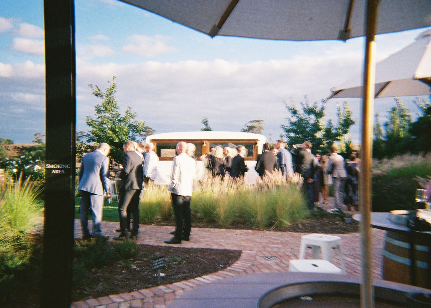 Wedding guests mingling outdoors near a vintage tram on a paved courtyard at golden hour.
