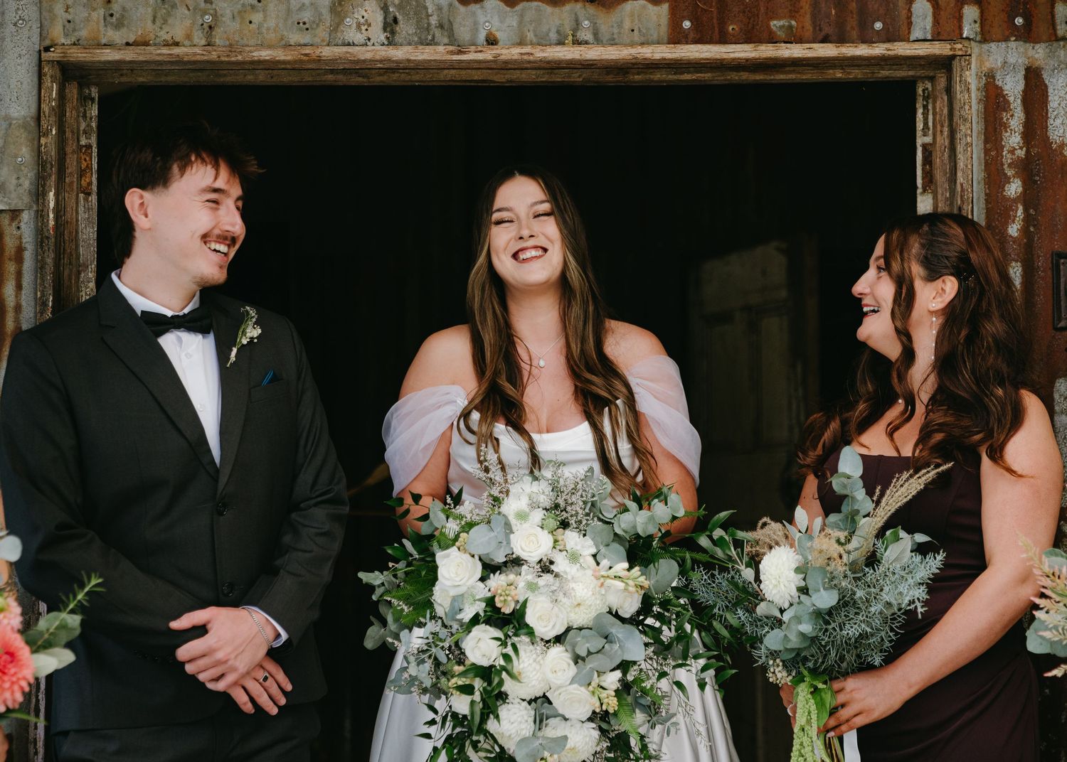 Bride laughing joyfully with groomsman and bridesmaid holding white floral bouquet in rustic barn.