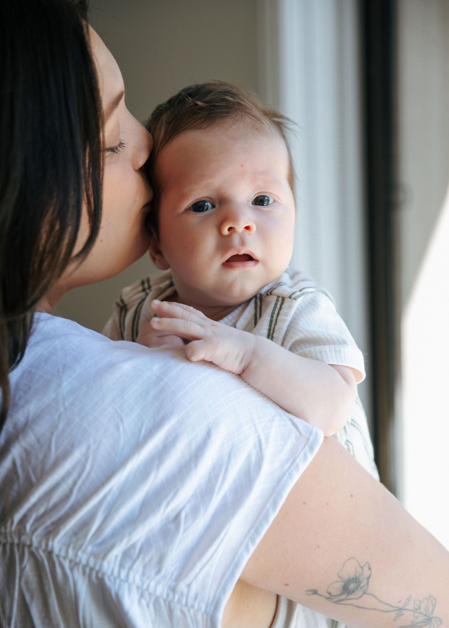 Mother kissing her alert newborn baby on the cheek near a bright window.