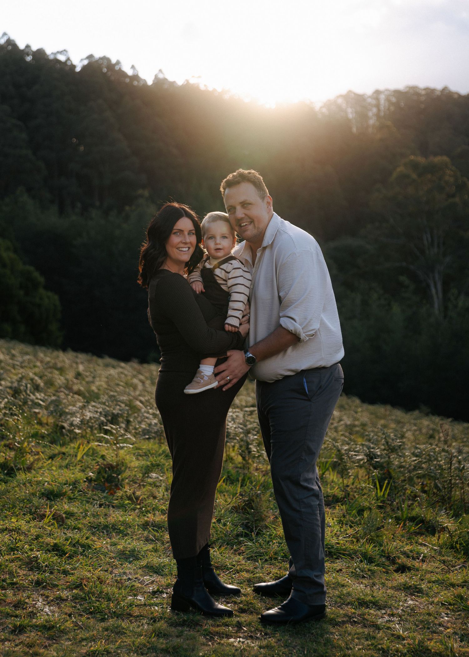 Smiling family with pregnant mom and dad holding toddler posing in golden sunset meadow.