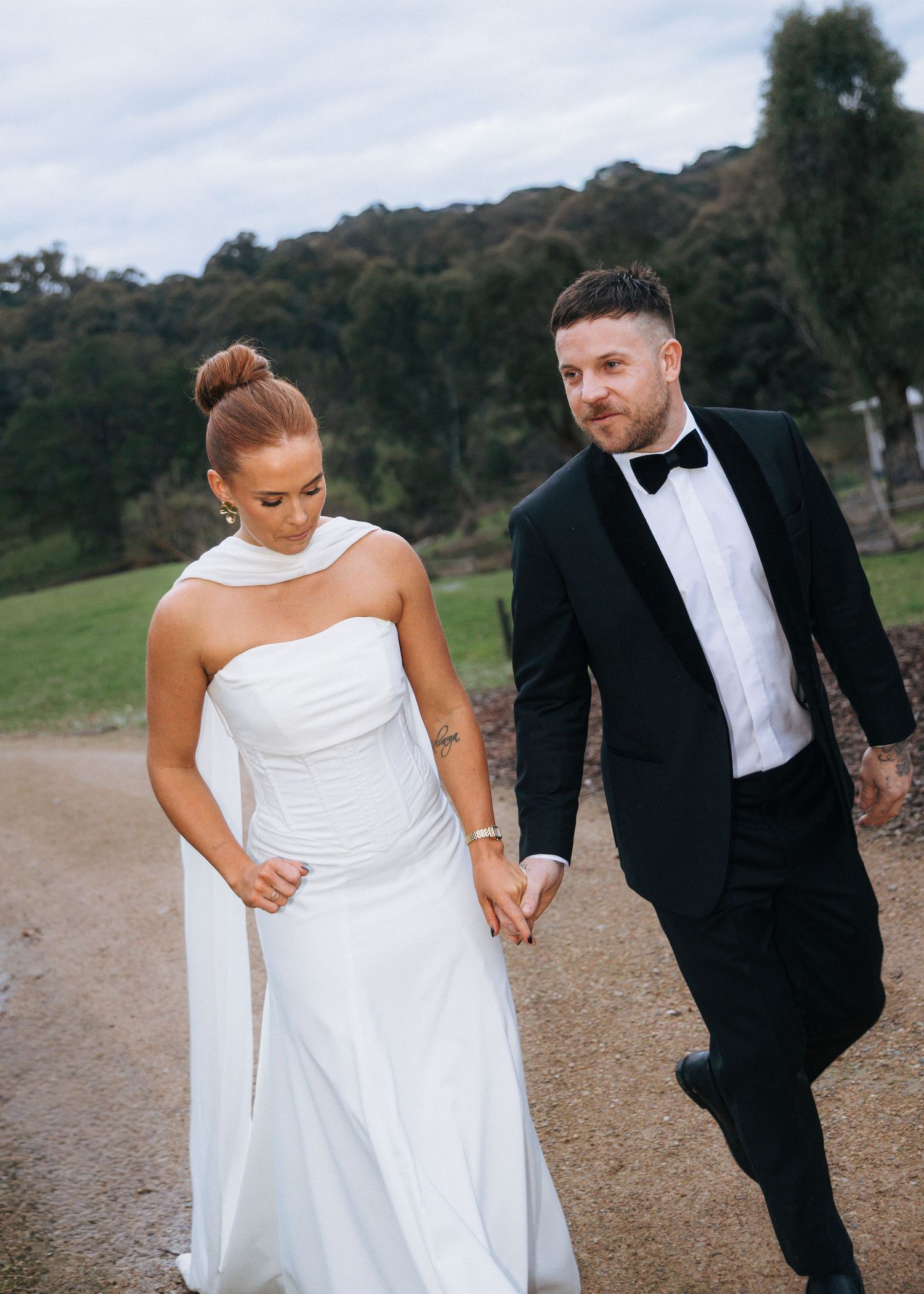 Bride in white gown and groom in black tuxedo walk hand in hand along a scenic outdoor path.