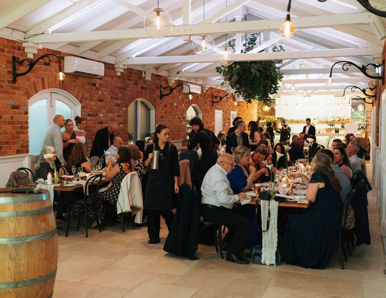 Wedding reception guests dining in a charming brick conservatory venue with warm lighting and rustic wine barrels.