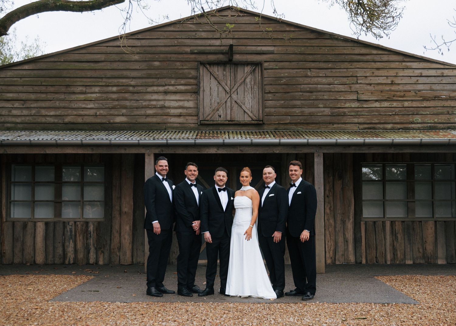 Wedding party poses in tuxedos and white gown before rustic wooden barn venue.