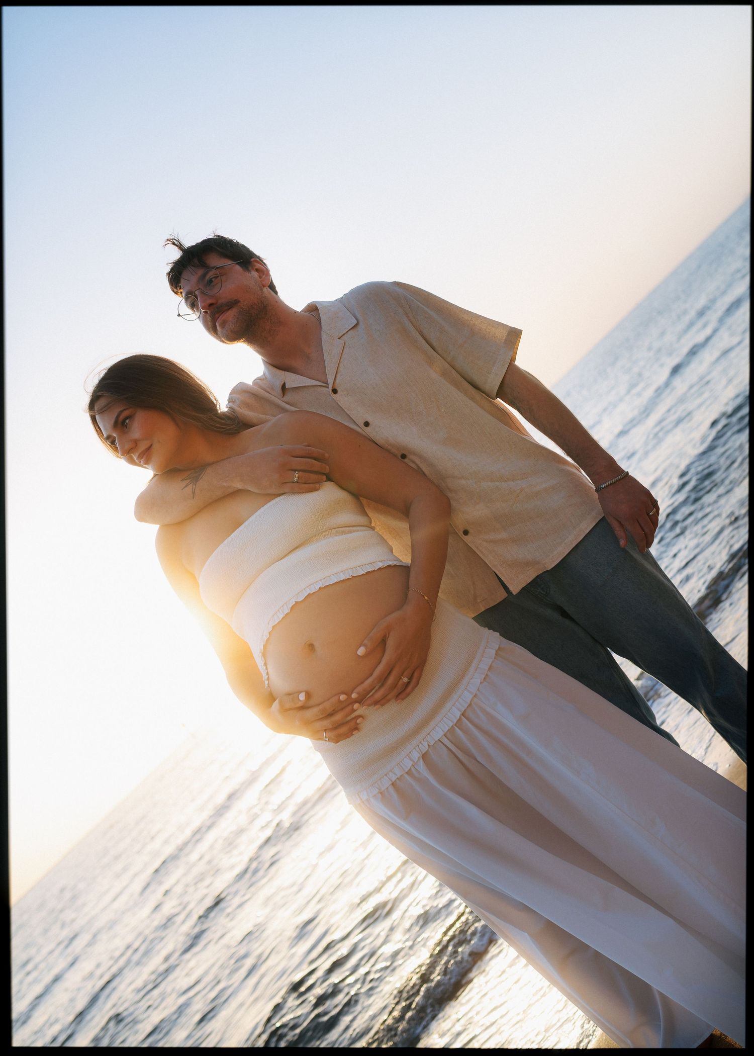 Backlit maternity photo of couple standing together on beach at golden hour by the ocean.