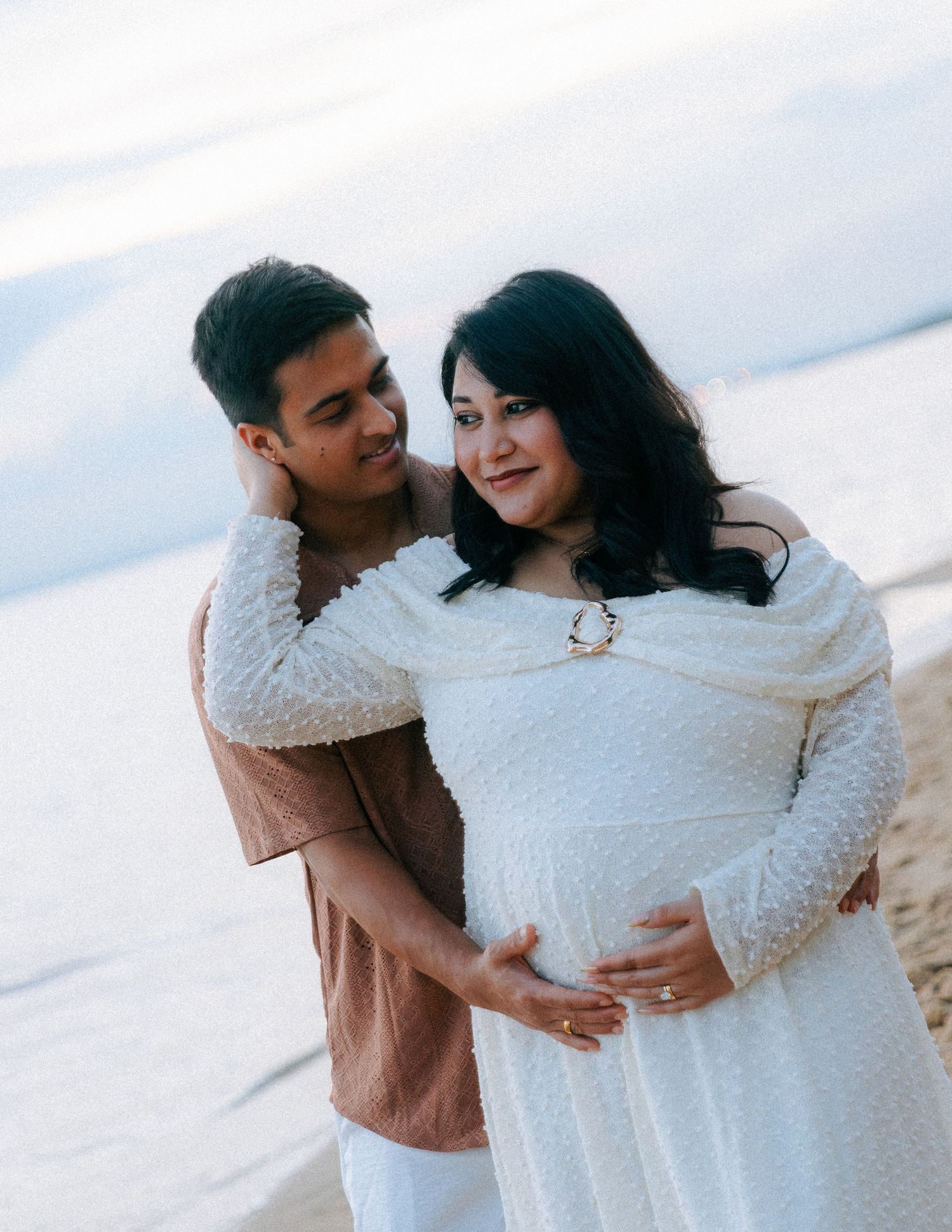 Couple maternity beach photo, man embracing pregnant woman in white lace dress at sunset.