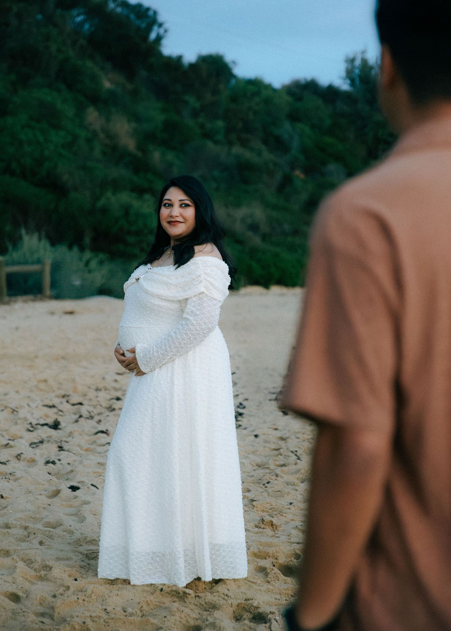 Pregnant woman in white off-shoulder dress smiling at partner on sandy beach at dusk.