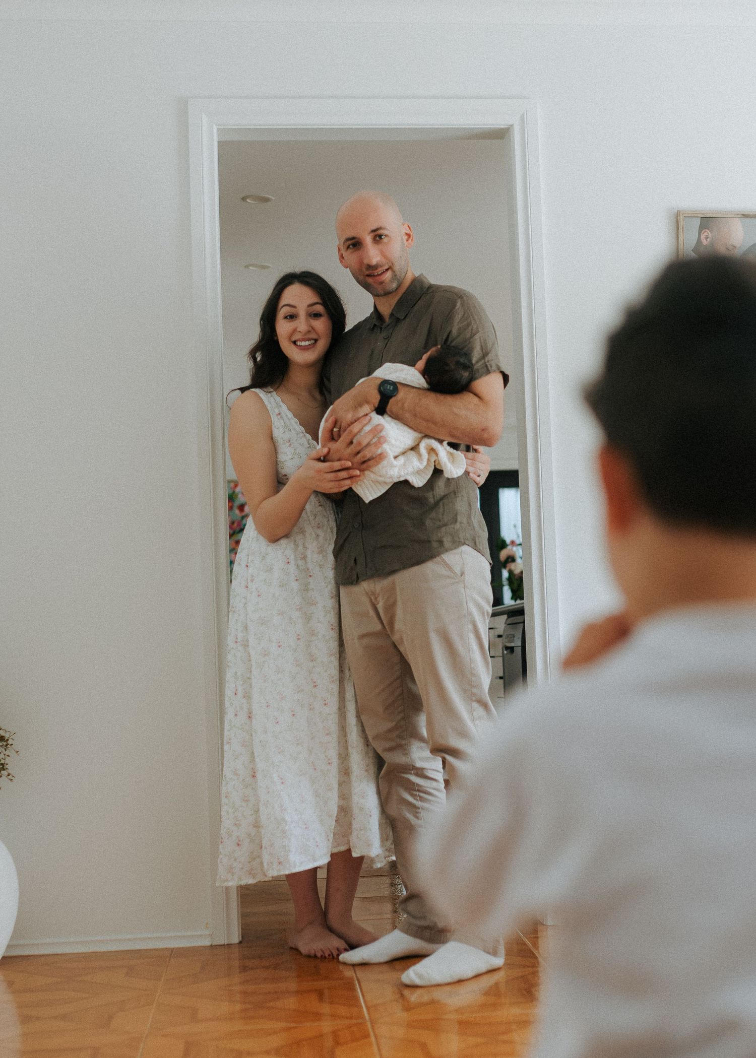 Couple standing in doorway holding newborn baby as toddler watches in bright home interior.