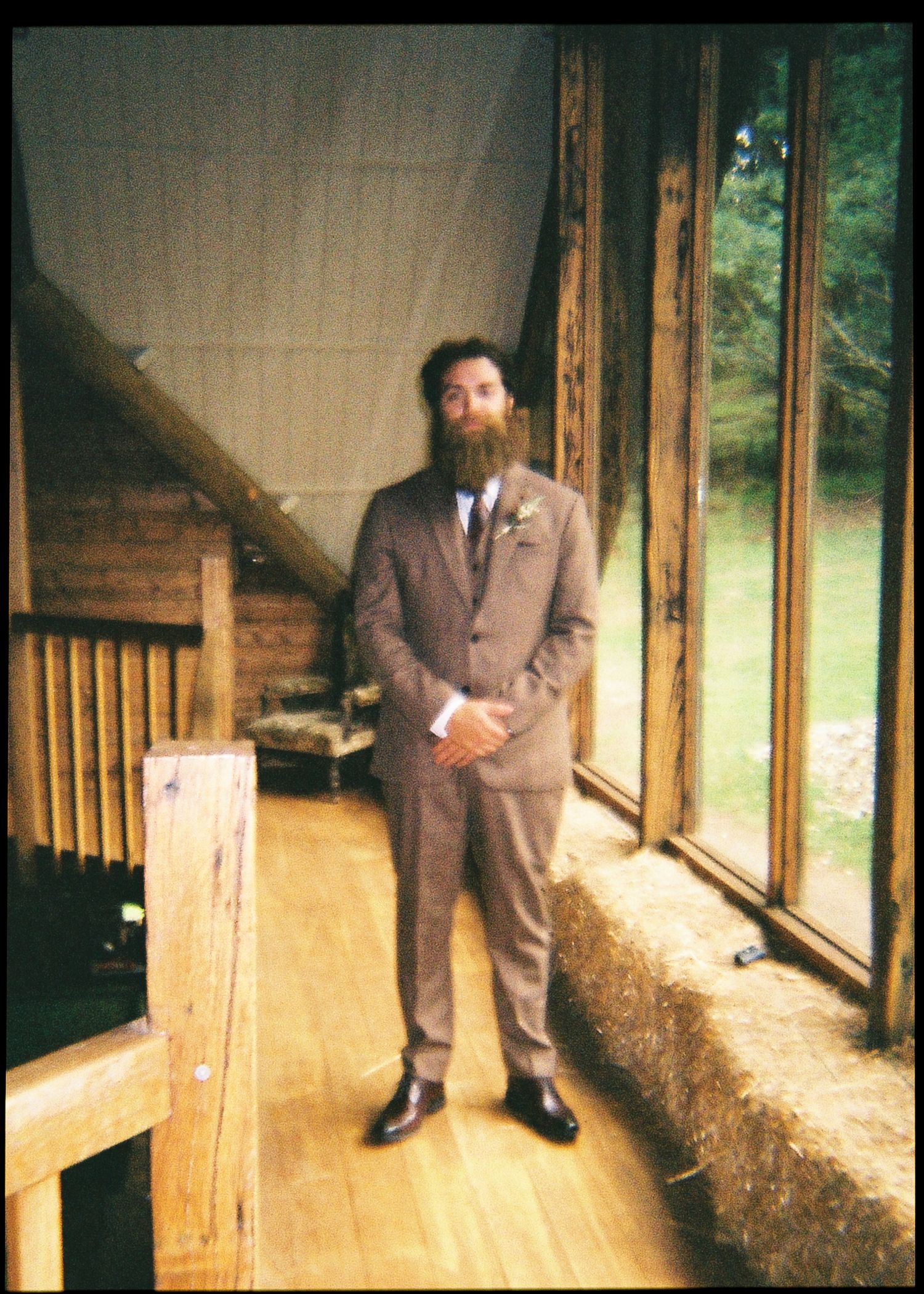 Bearded groom in brown suit with boutonniere standing in rustic wooden cabin venue.