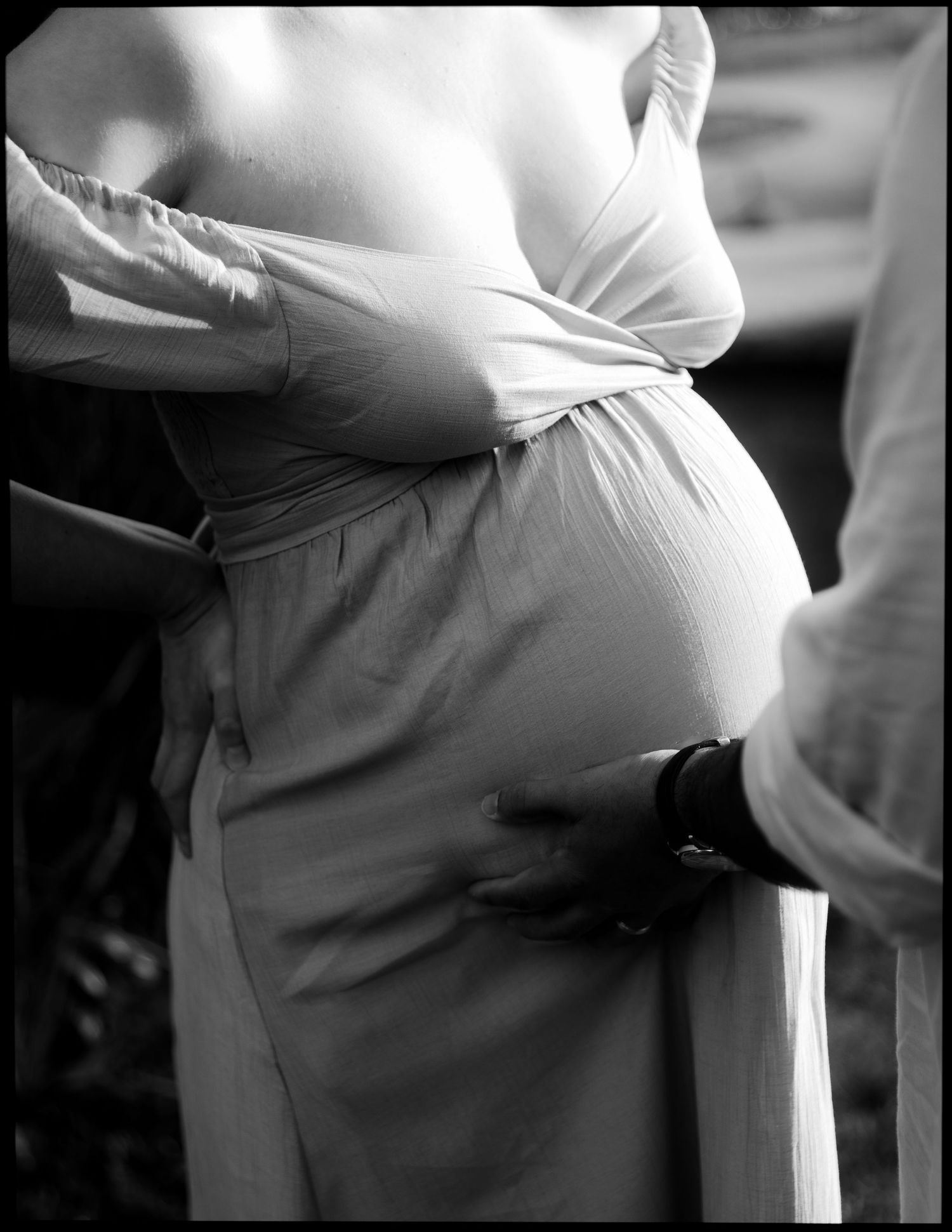 Black and white close-up of pregnant belly in flowing dress with partner tenderly placing hand on back.