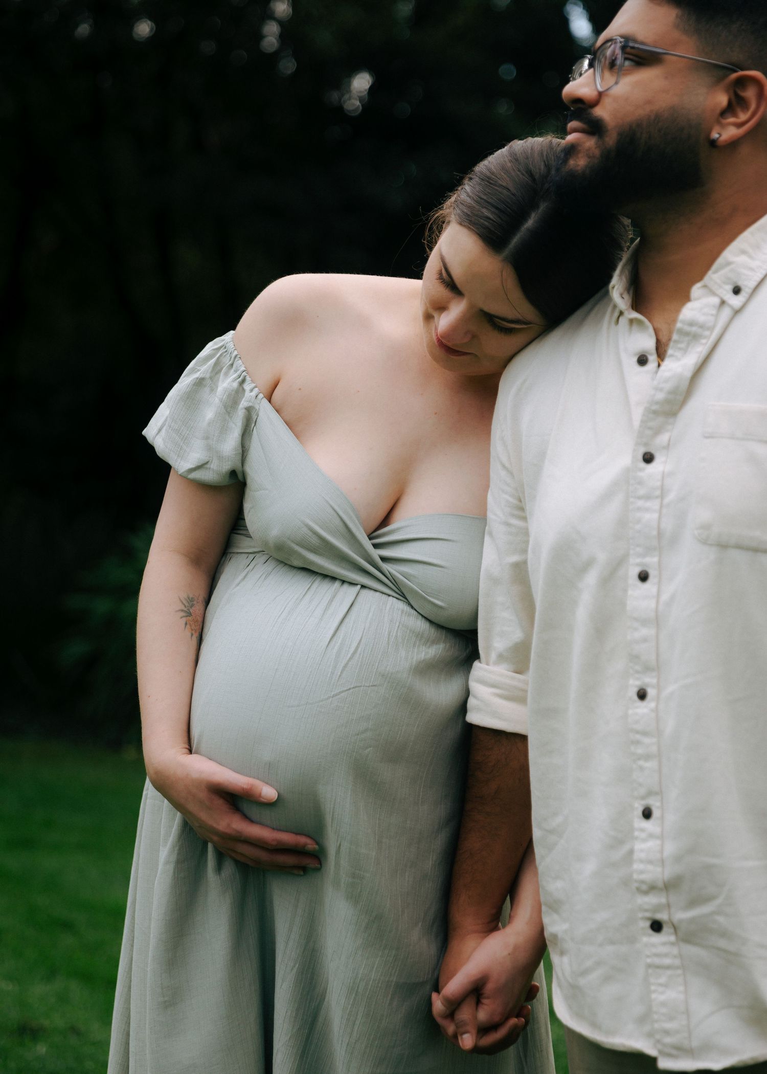 Expecting couple standing close together in garden, woman in sage maternity dress resting on partner.