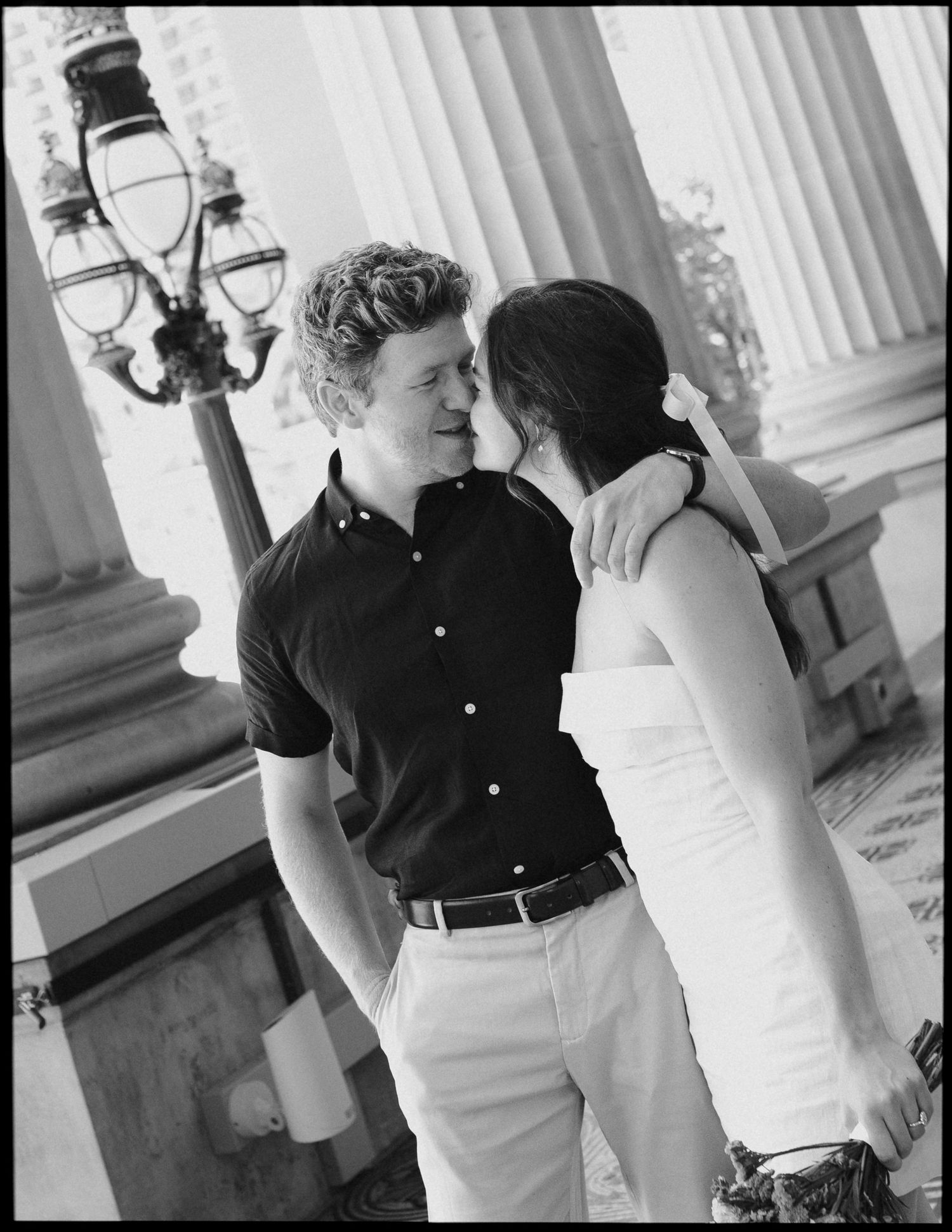 Black and white photo of a couple sharing a romantic moment beside classical columns at a historic venue.