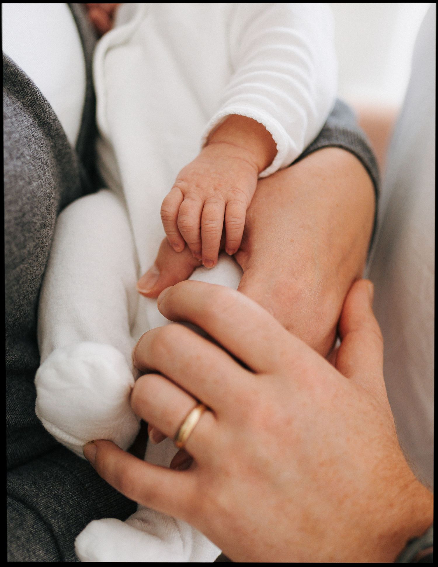 Close-up of a newborn baby's tiny hand resting on an adult's hand wearing a gold wedding ring, with the baby in soft white clothing.