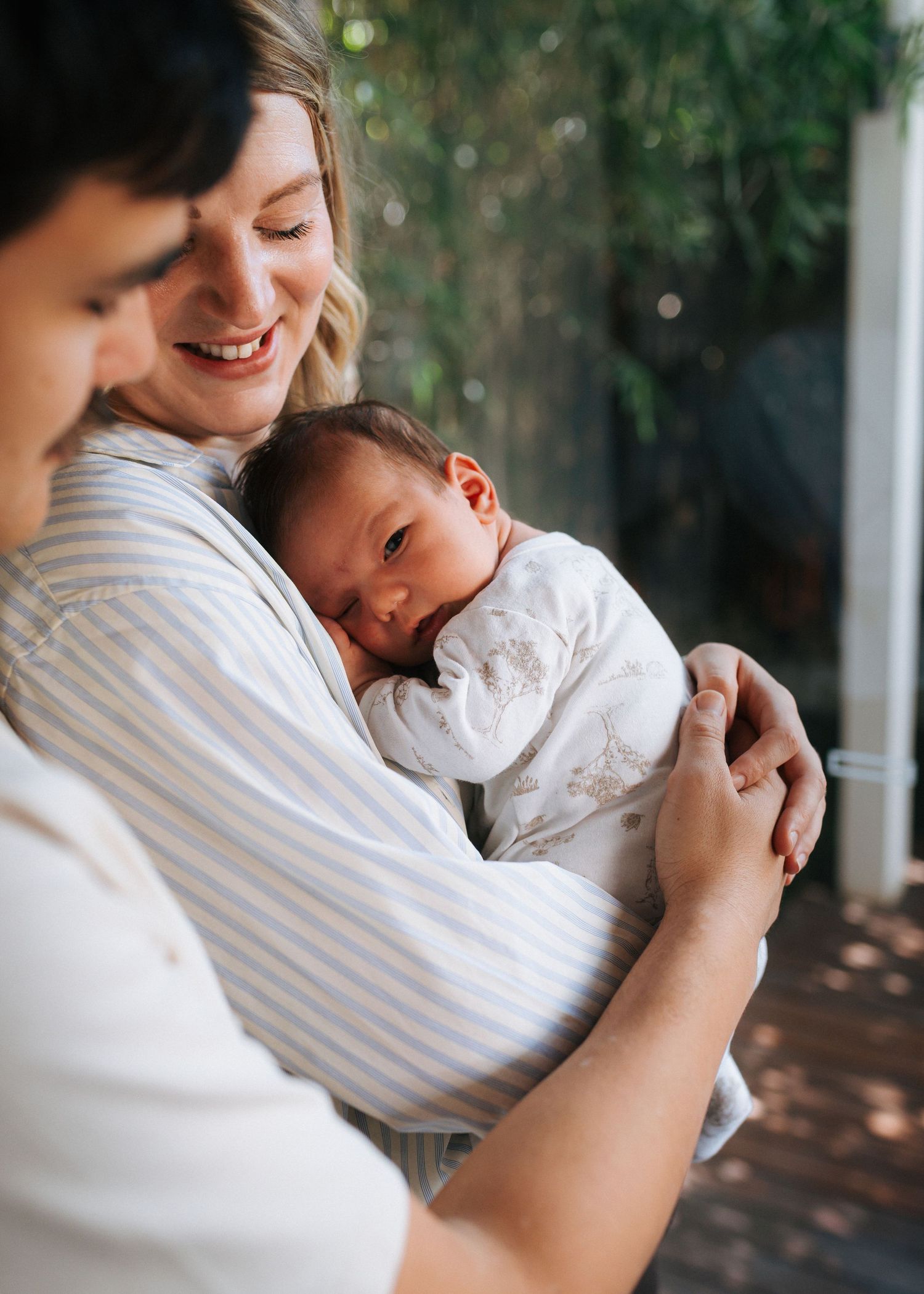 A smiling woman in a striped shirt cradles a sleeping newborn baby in white clothing while a man embraces them outdoors.