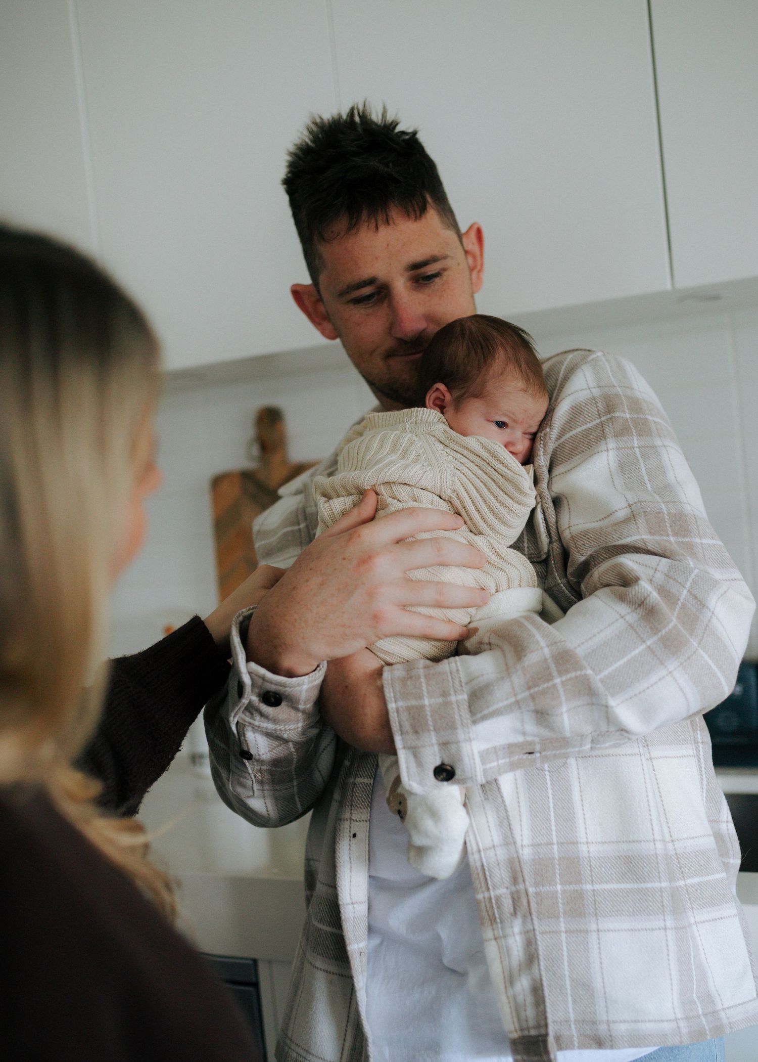 Father in plaid shirt cradling sleeping newborn baby in bright kitchen interior.